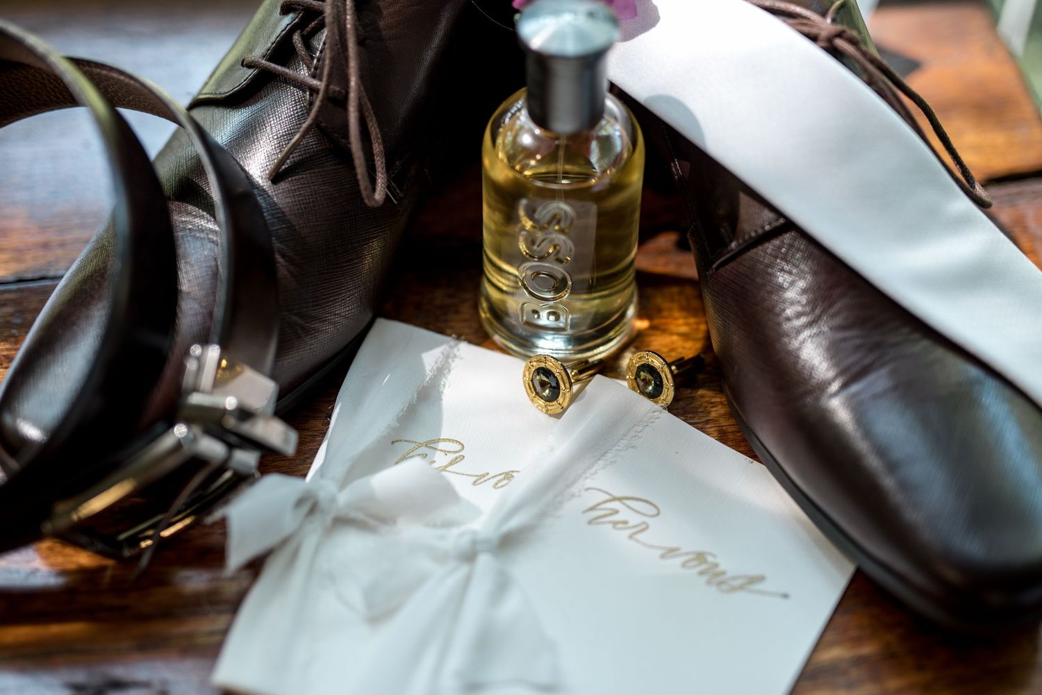 Close-up of groom wedding details including cologne bottle, leather shoes, cufflinks, and vow card.