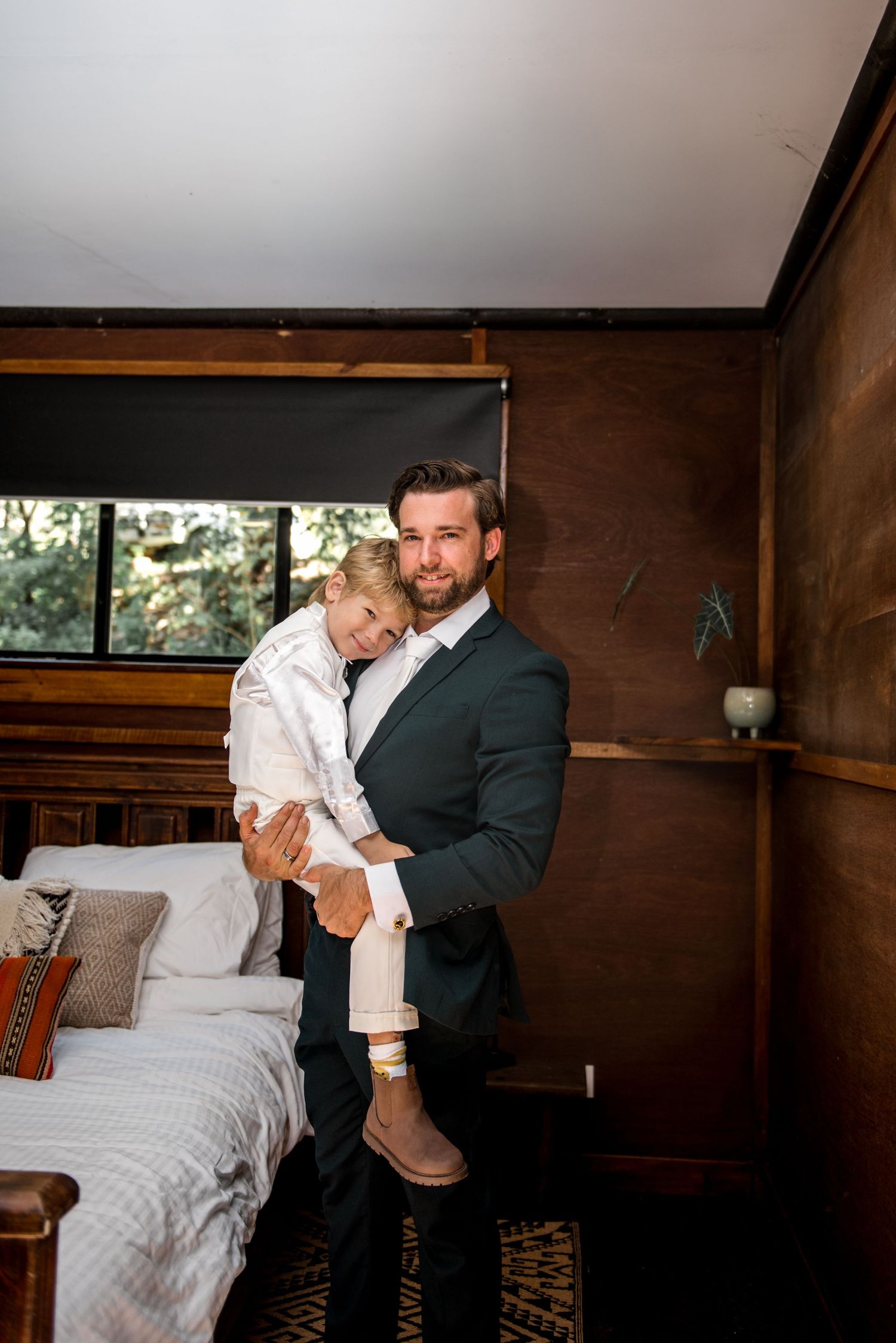 Groom in dark suit tenderly holds a young blonde boy in white outfit in a wooden cabin bedroom.