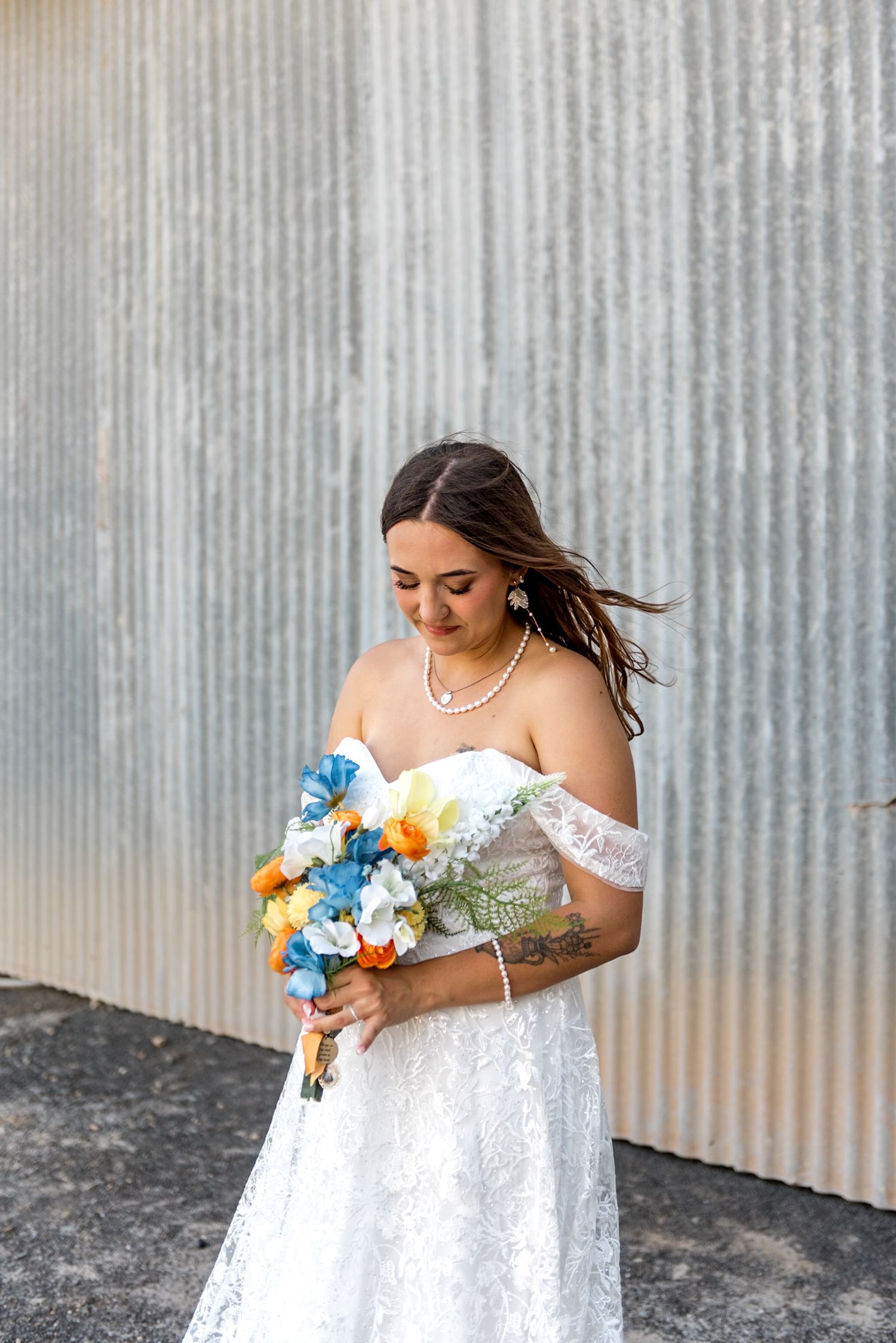 Bride in off-shoulder lace gown looks down at vibrant blue and orange bouquet, standing before corrugated metal wall.