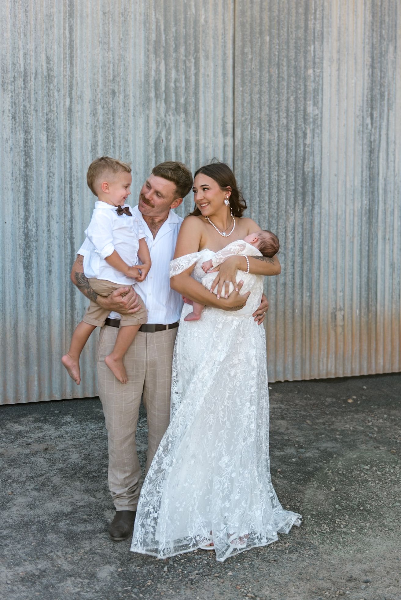 Happy wedding family laughing together, bride holding newborn baby and groom lifting toddler boy outdoors.