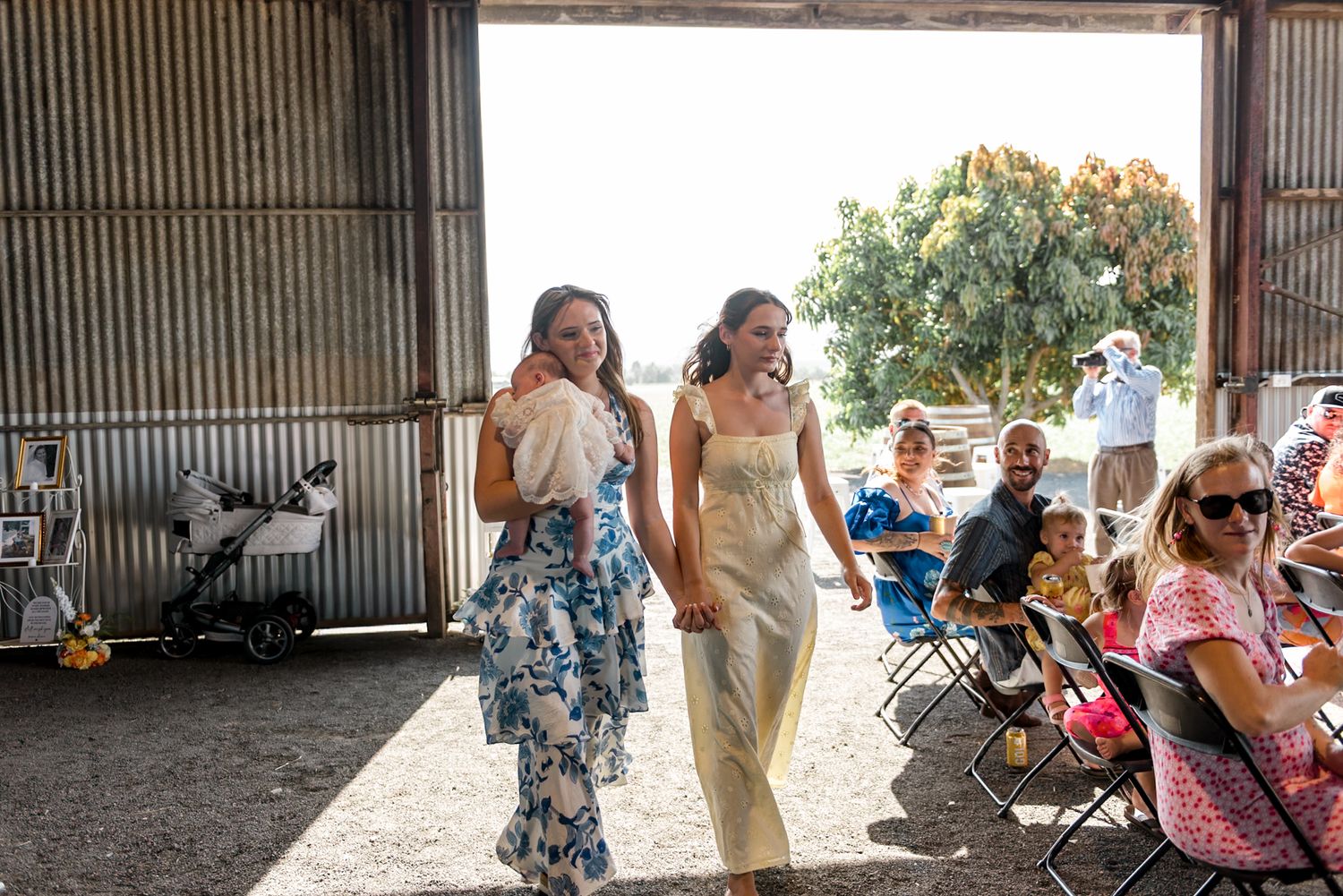 Two bridesmaids in floral and yellow dresses walk down the aisle inside a rustic barn wedding ceremony.