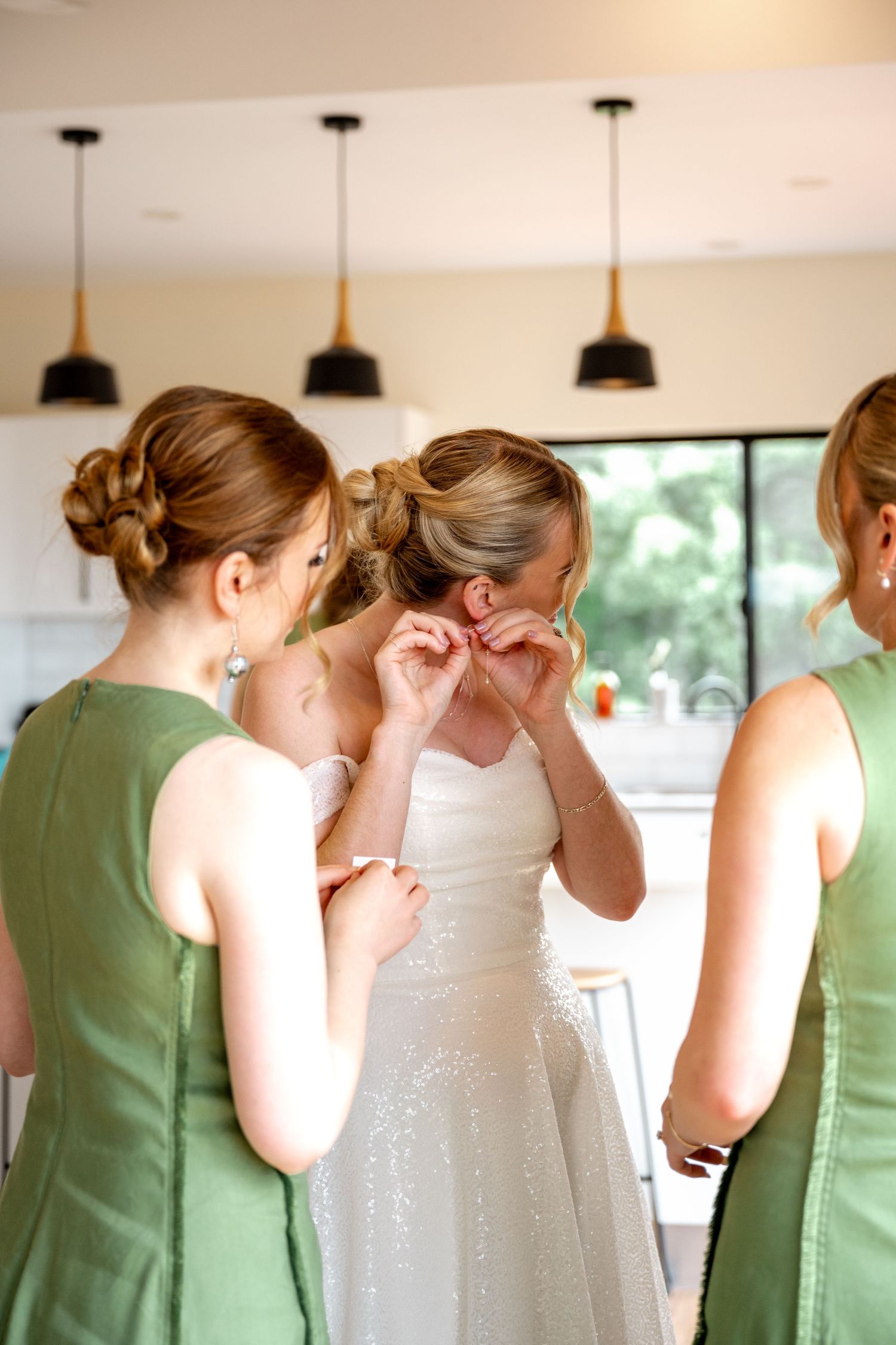 Bridesmaids in green dresses assist bride with earrings during wedding morning preparations.