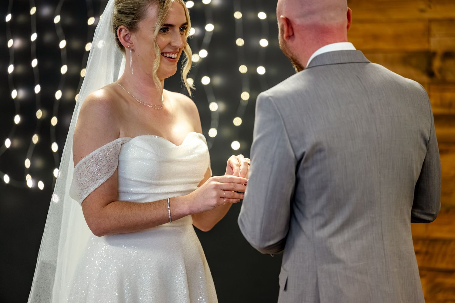 Bride in white gown and veil smiling while placing ring on groom during wedding ceremony.