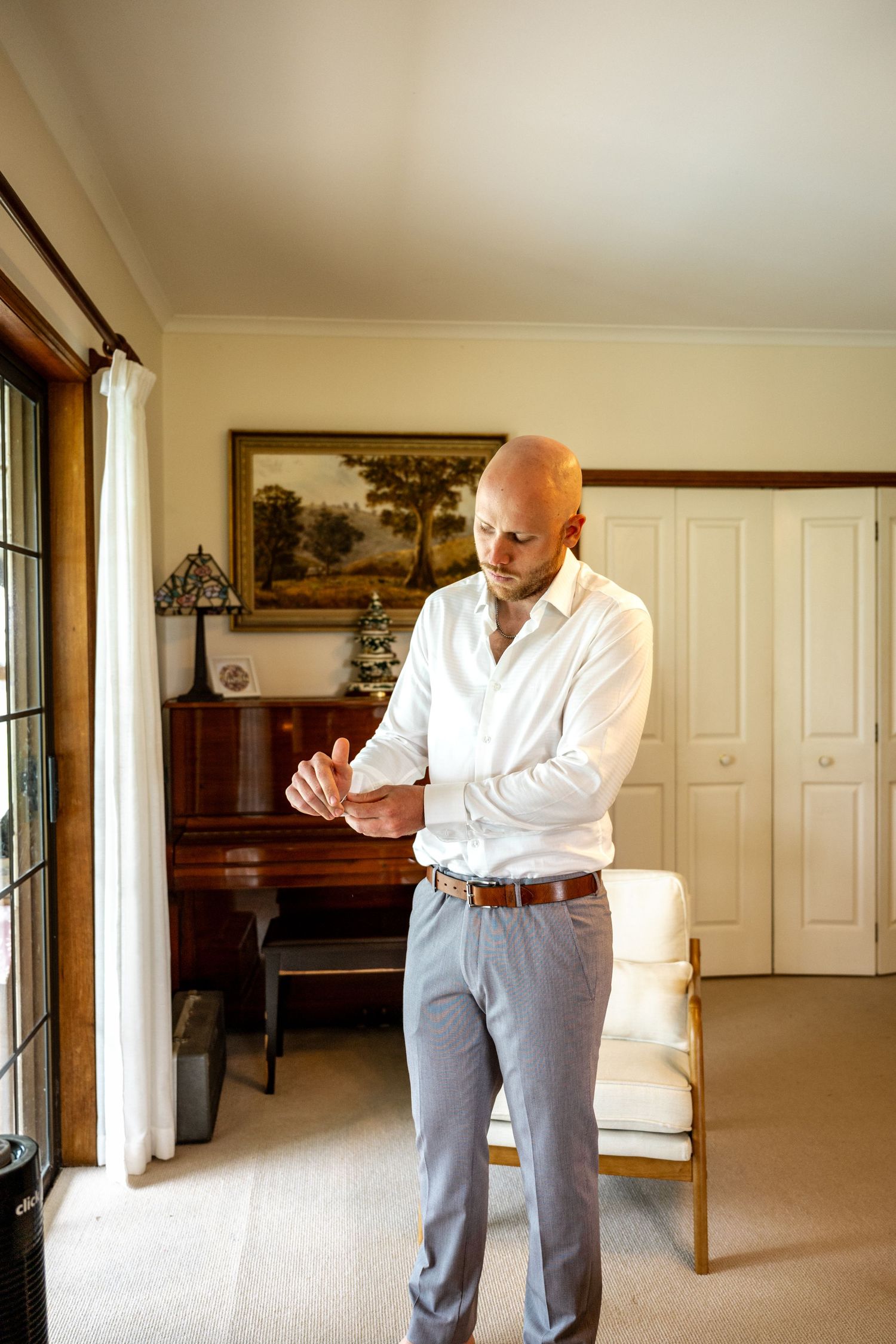 Groom in white shirt adjusting cufflinks in elegant room with vintage piano backdrop.