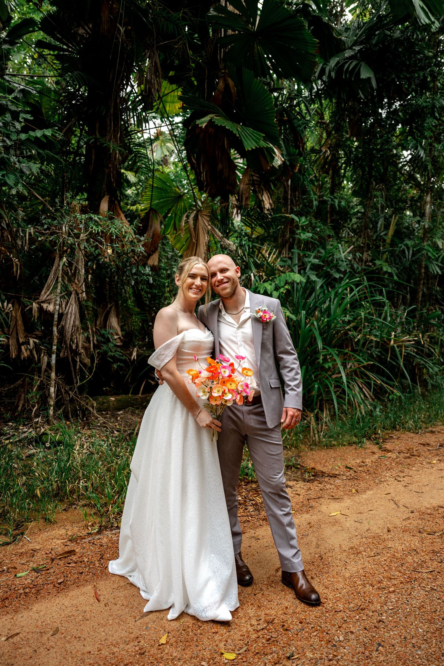 Smiling wedding couple pose together on a tropical dirt path surrounded by lush green jungle foliage.