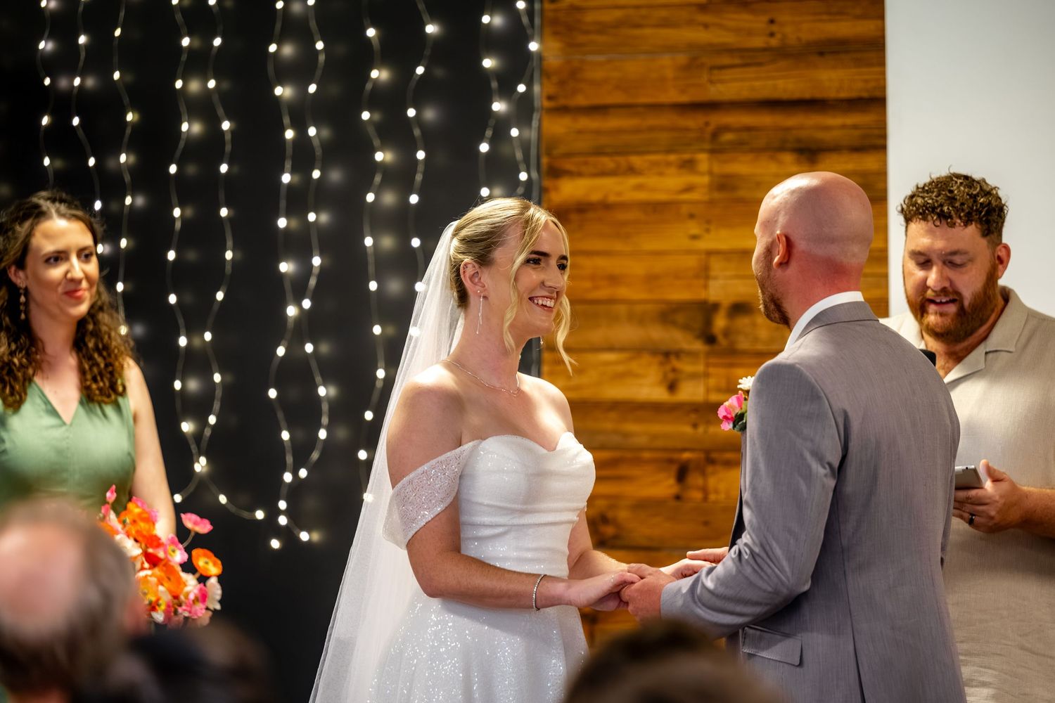 Radiant bride in white veil smiles at groom while holding hands during wedding ceremony with fairy lights.