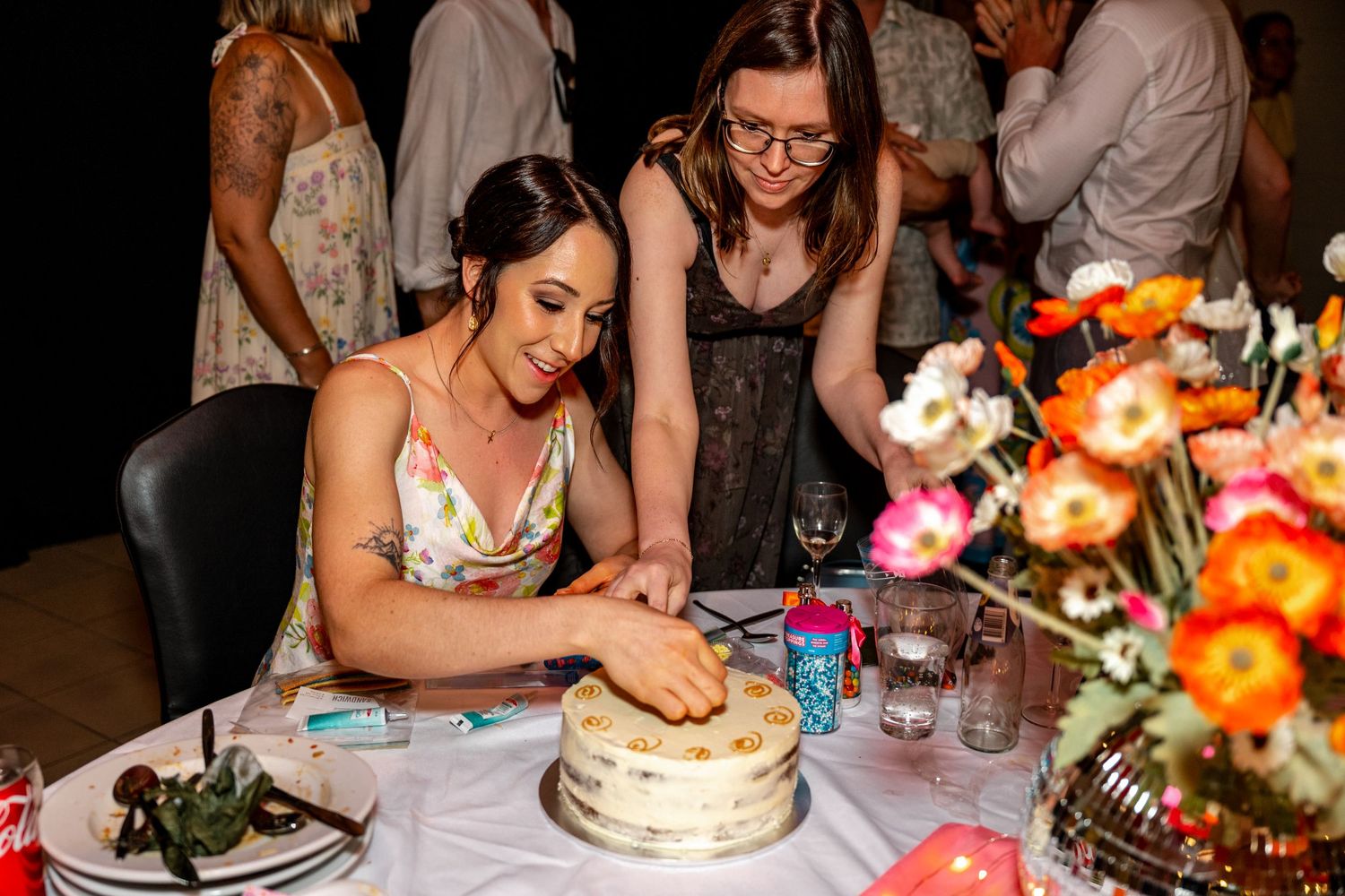 Woman in floral dress cutting white wedding cake at decorated reception table with colorful flowers.