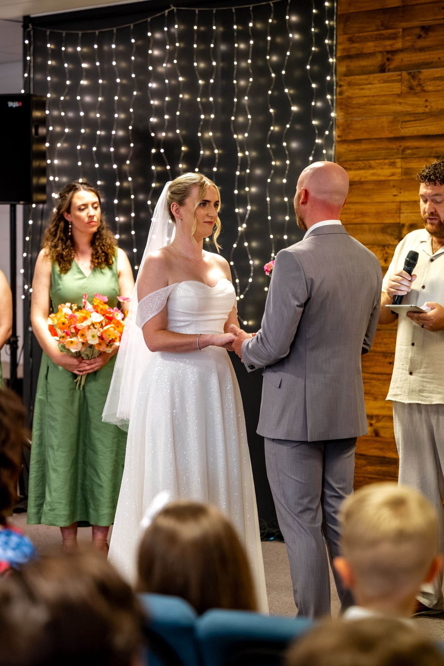 Bride in white off-shoulder gown holds hands with groom at altar with fairy light backdrop and bridesmaid beside her.