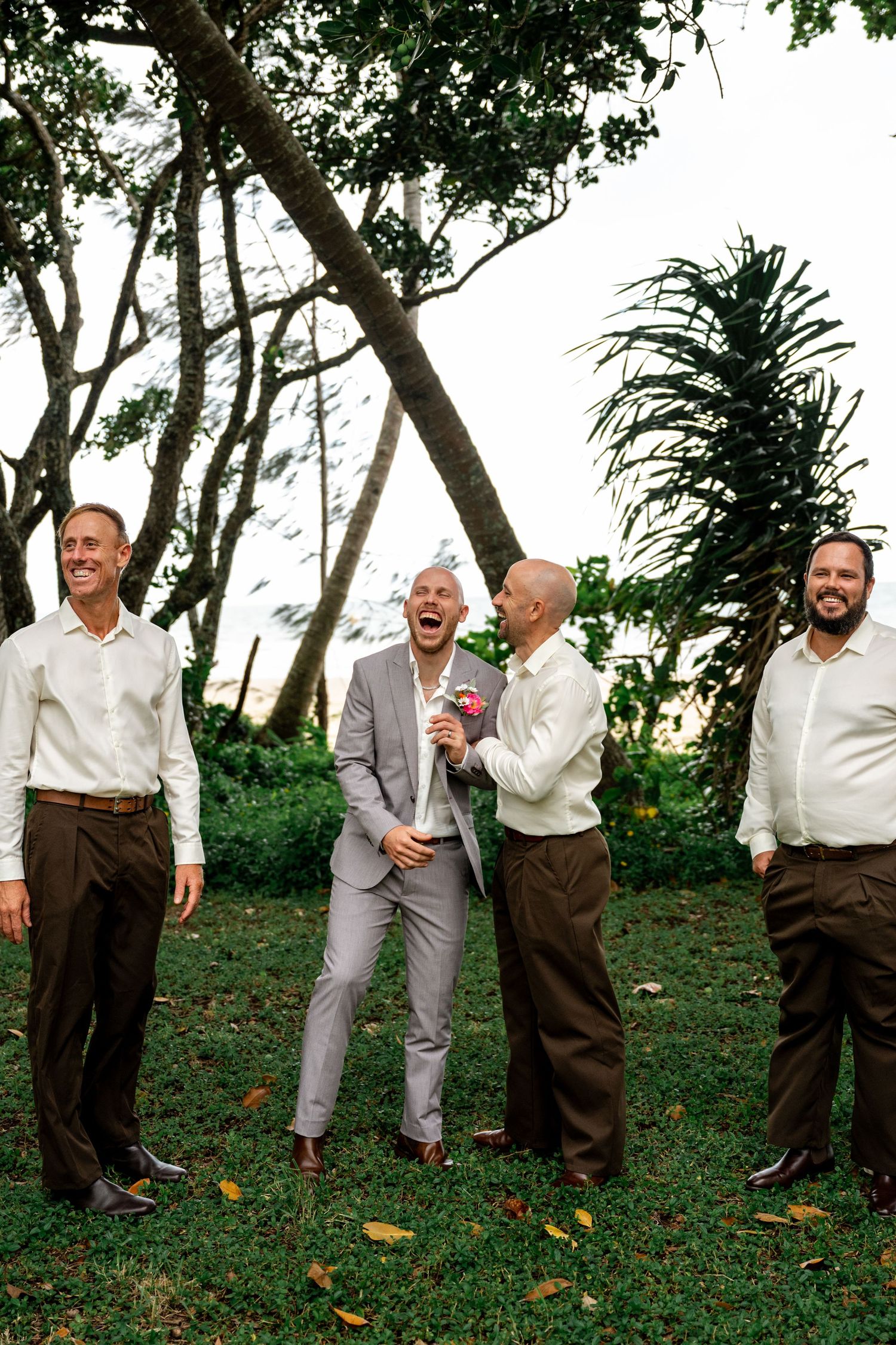Groom in grey suit laughs heartily with groomsmen in white shirts and brown pants in lush tropical garden setting.