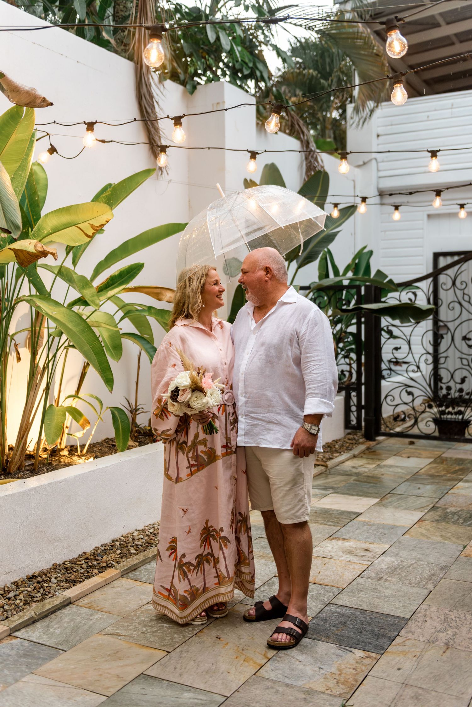 Bride and groom sharing romantic moment under clear umbrella in tropical courtyard with festoon lights.