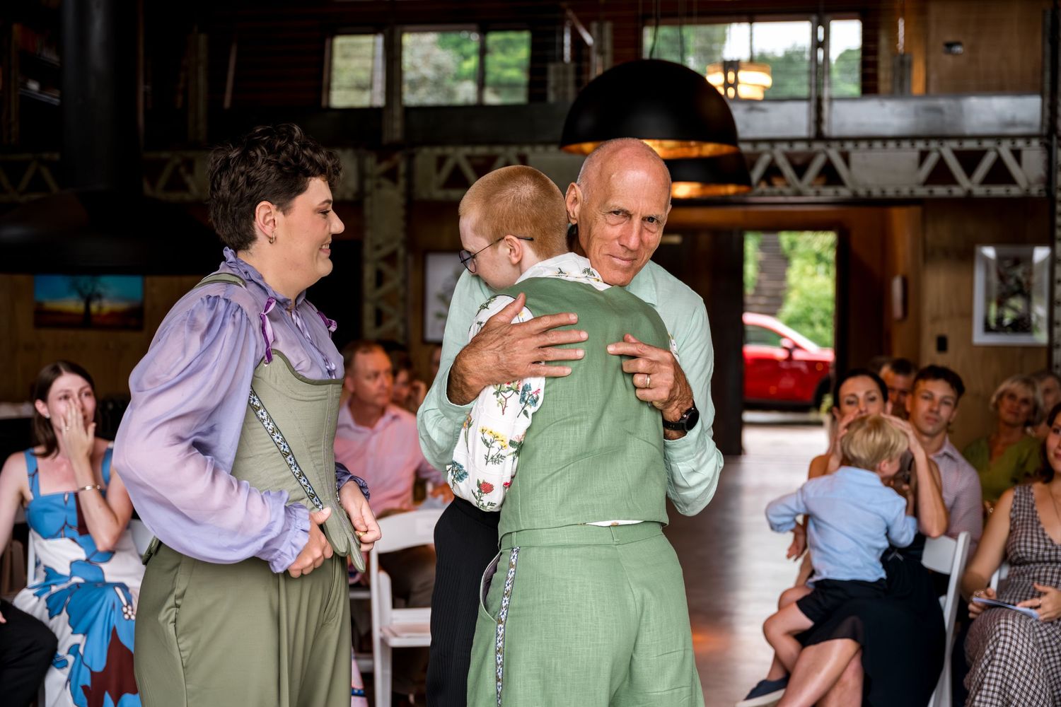 Older man embraces bride in green overalls while another bride looks on at wedding ceremony.