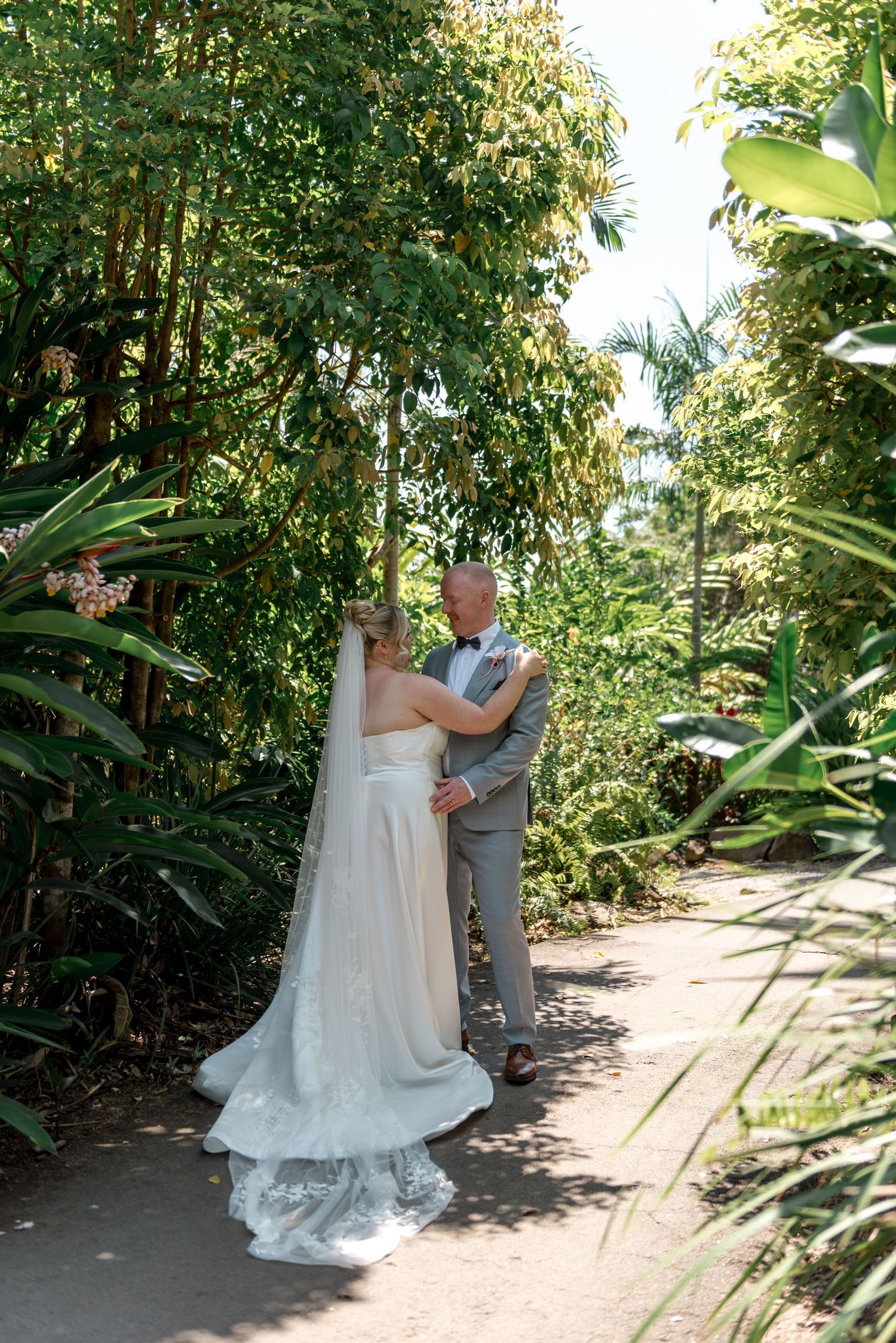 Bride and groom share a tender embrace on a shaded garden path surrounded by lush tropical greenery.