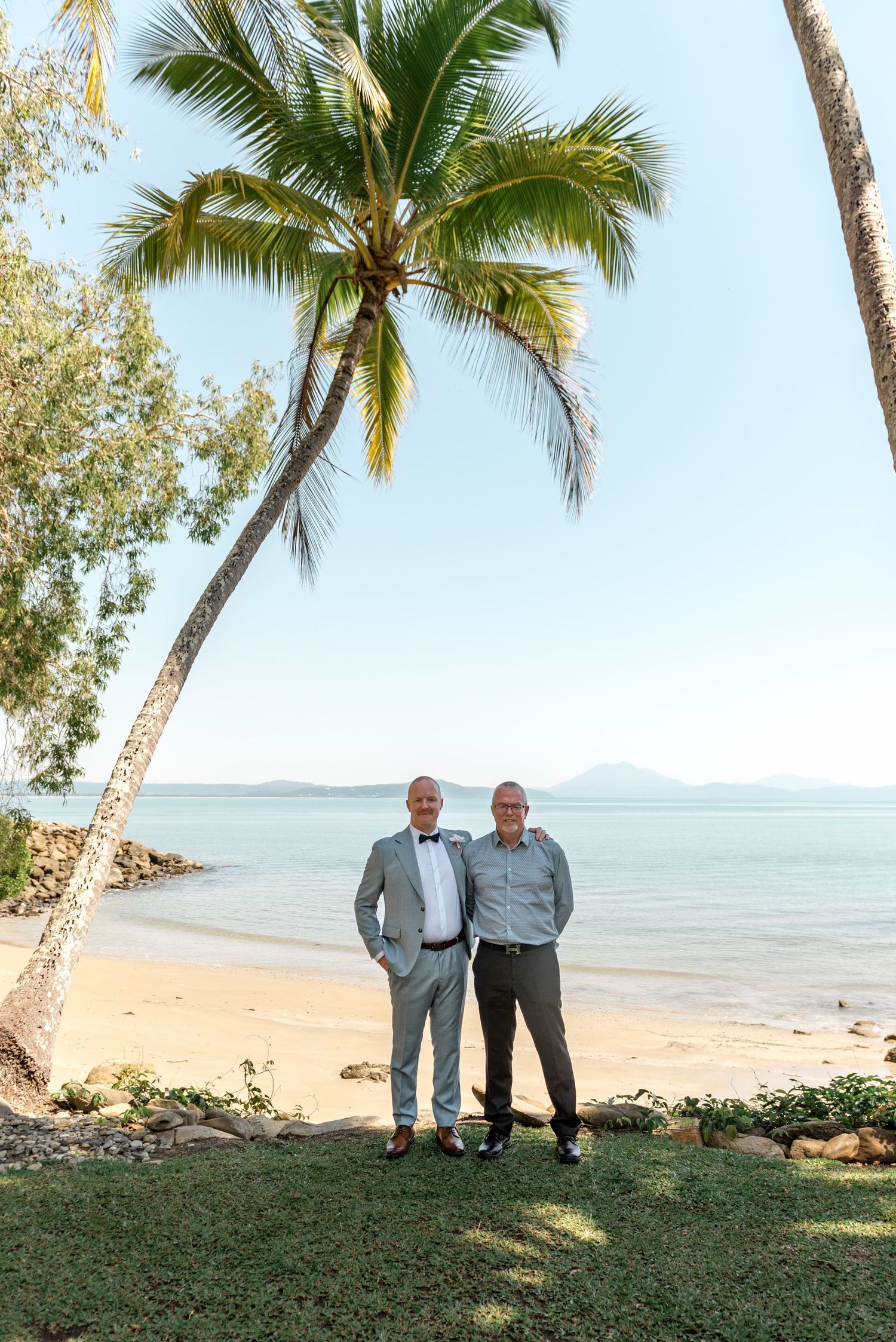 Two men in formal attire stand together under tall palm trees on a tropical sandy beach.