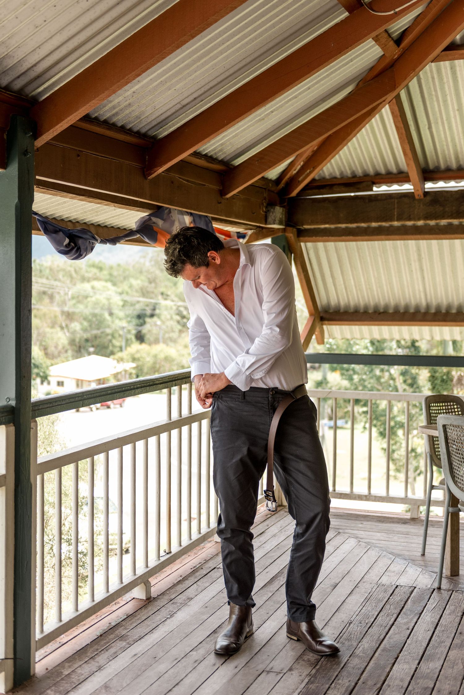 Groom in white shirt adjusting his suspenders while getting ready on a rustic outdoor balcony.