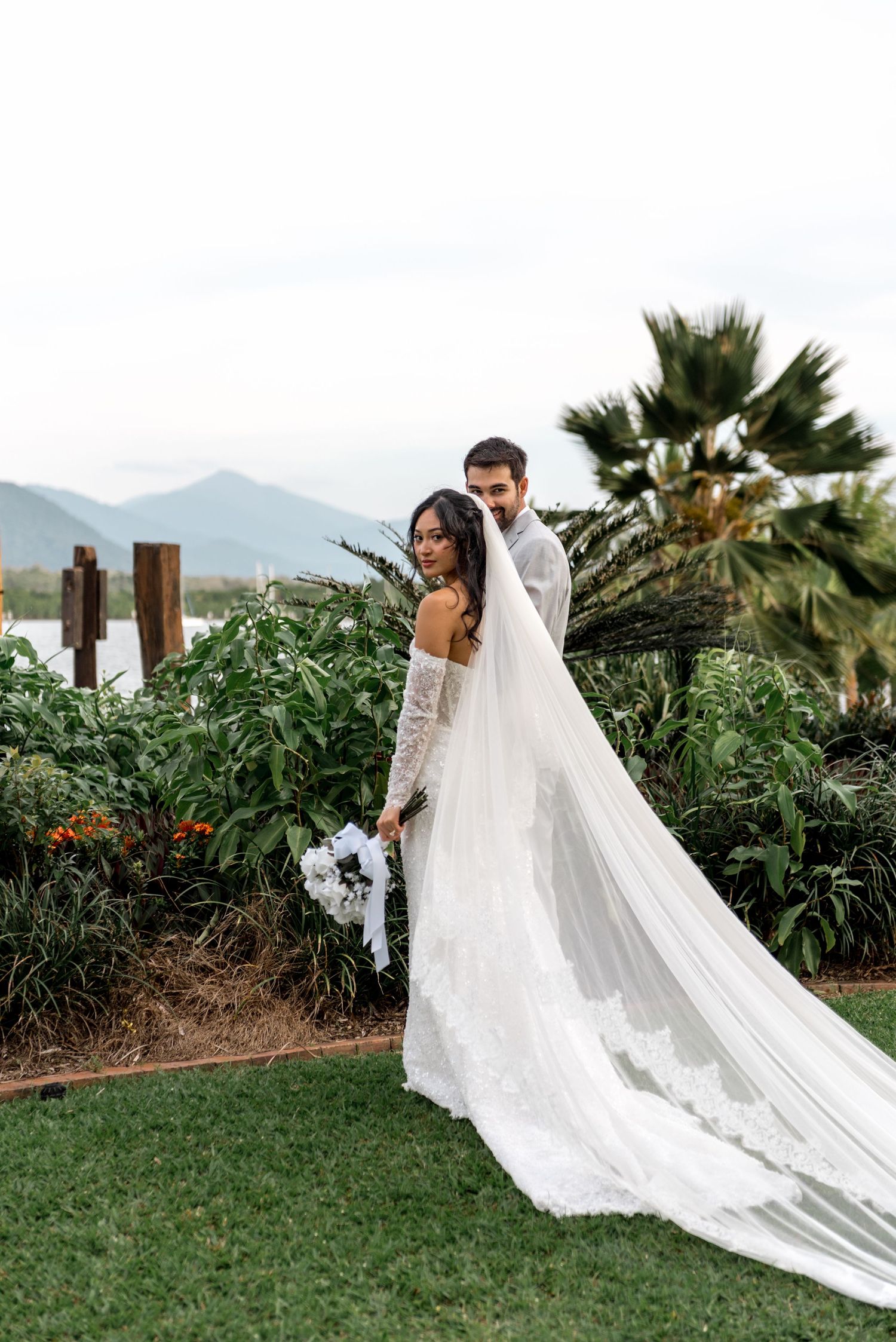 Bride and groom pose in tropical garden with mountains backdrop, bride wearing flowing lace veil.