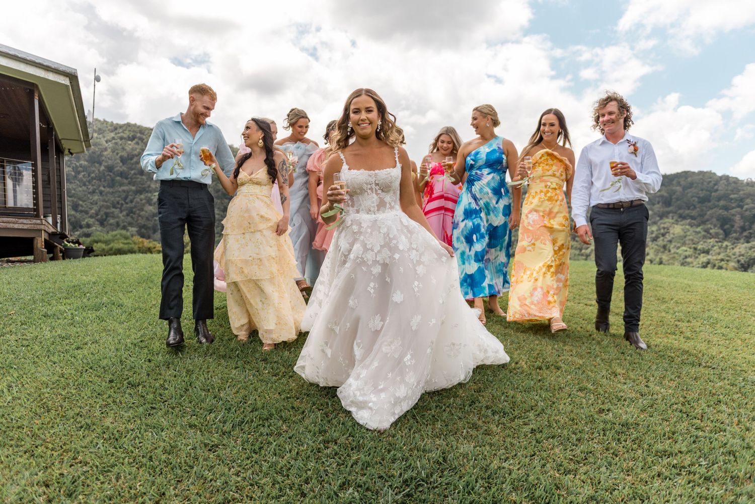 Radiant bride leads smiling wedding guests across green lawn with scenic mountain background.