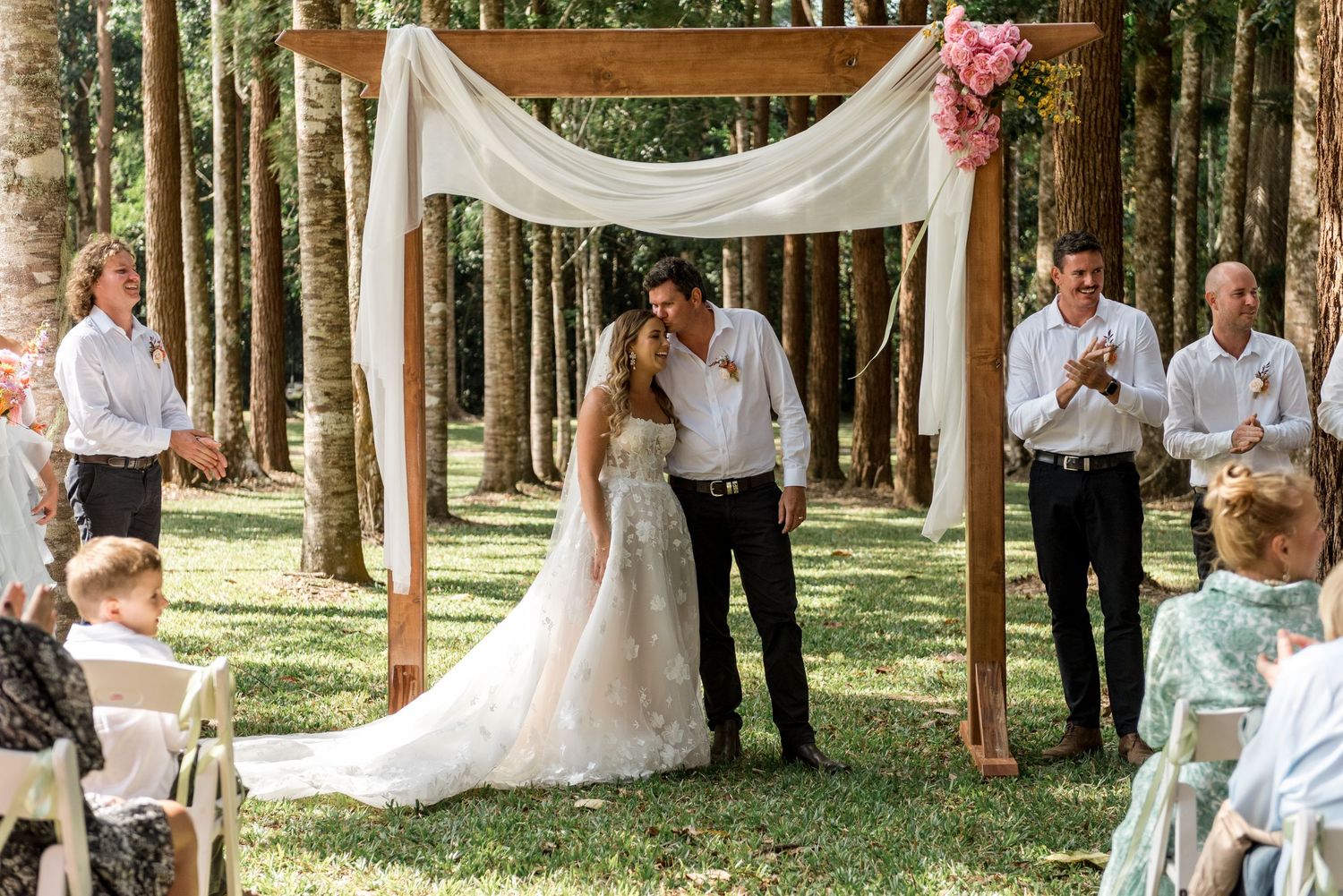 Newlyweds kiss under a rustic wooden arch with white fabric and floral decor as guests applaud in a woodland setting.