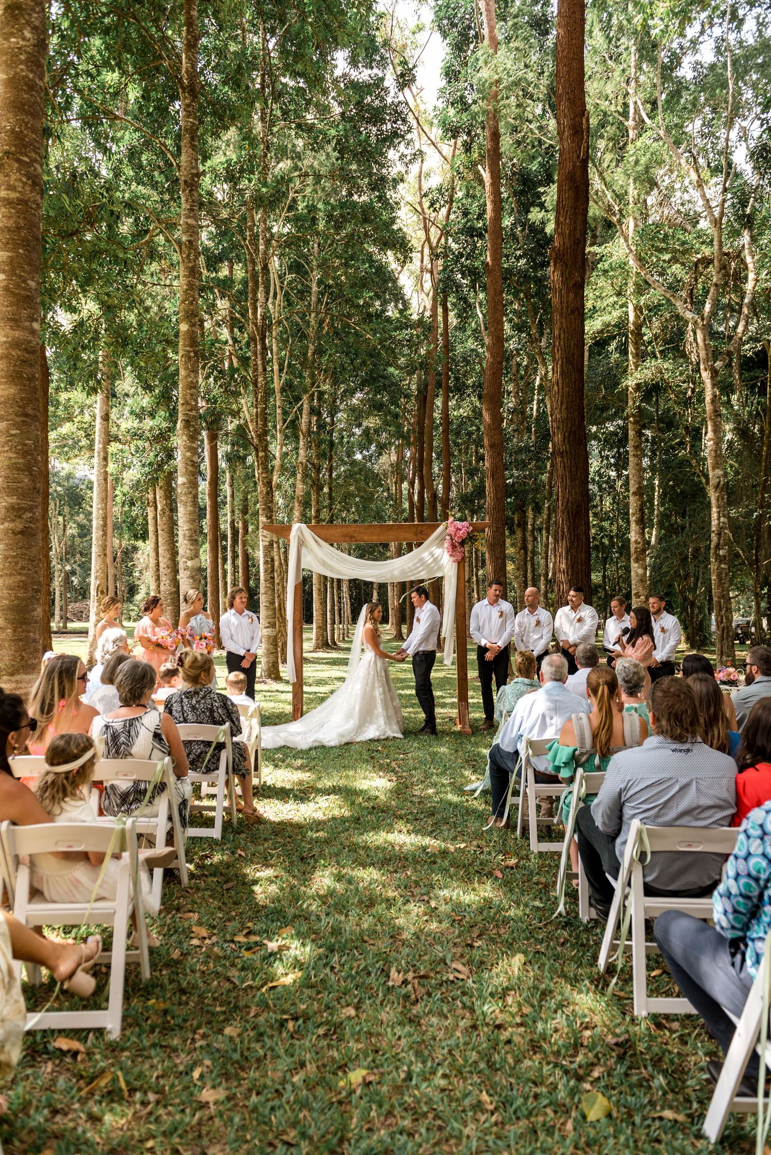 Wide view of an outdoor forest wedding ceremony with guests seated along a grass aisle.