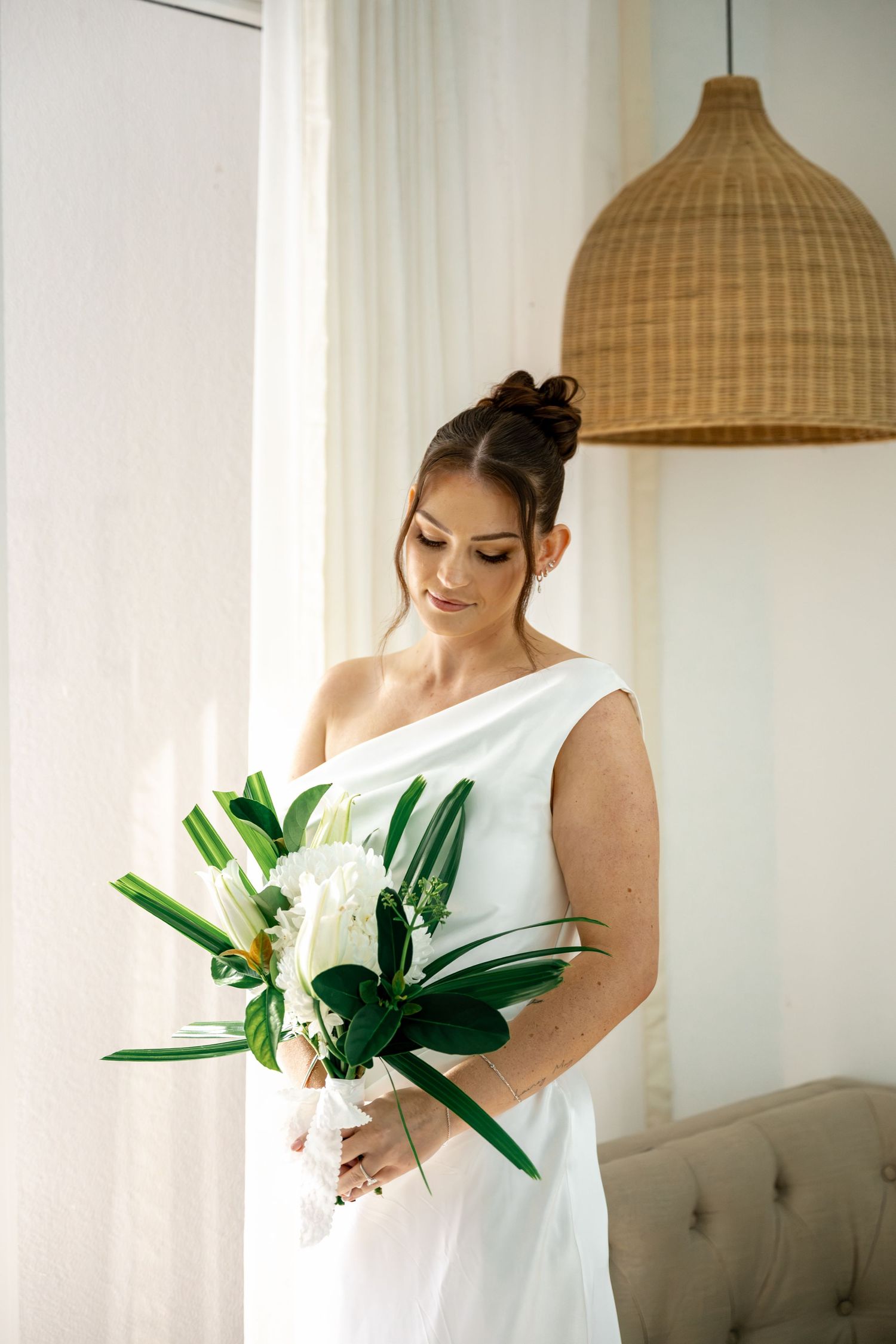Bride in white one-shoulder dress gazing down at tropical green and white wedding bouquet by curtained window at Sheraton Mirage Resort.