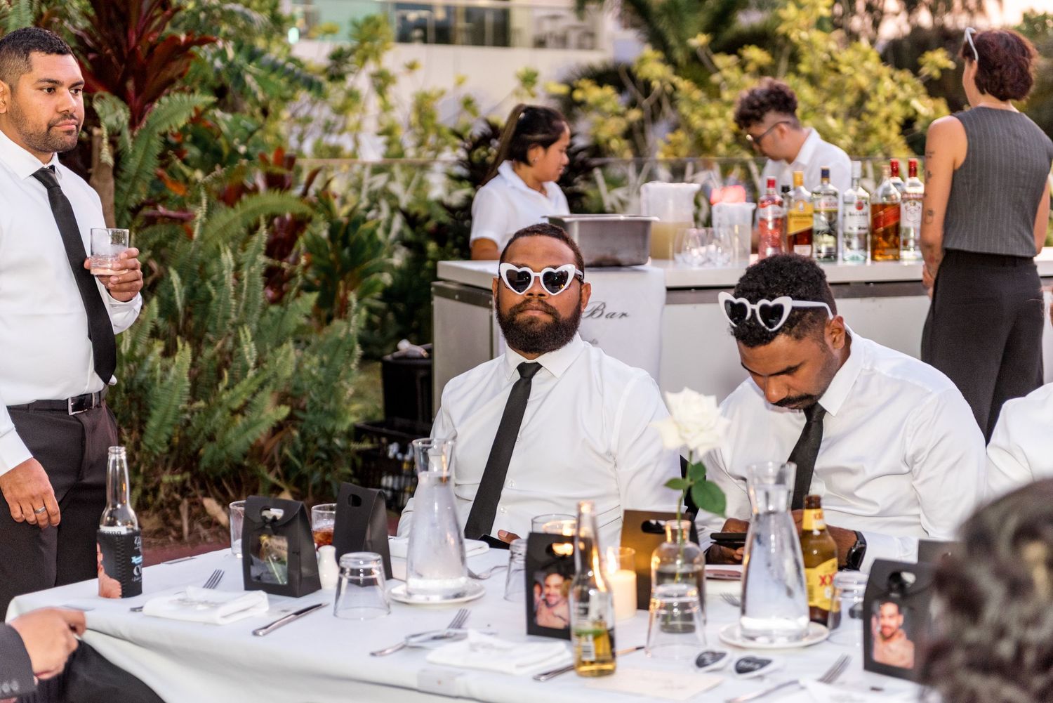 Two male wedding guests wearing novelty heart-shaped sunglasses sit at a reception table with drinks and candles.