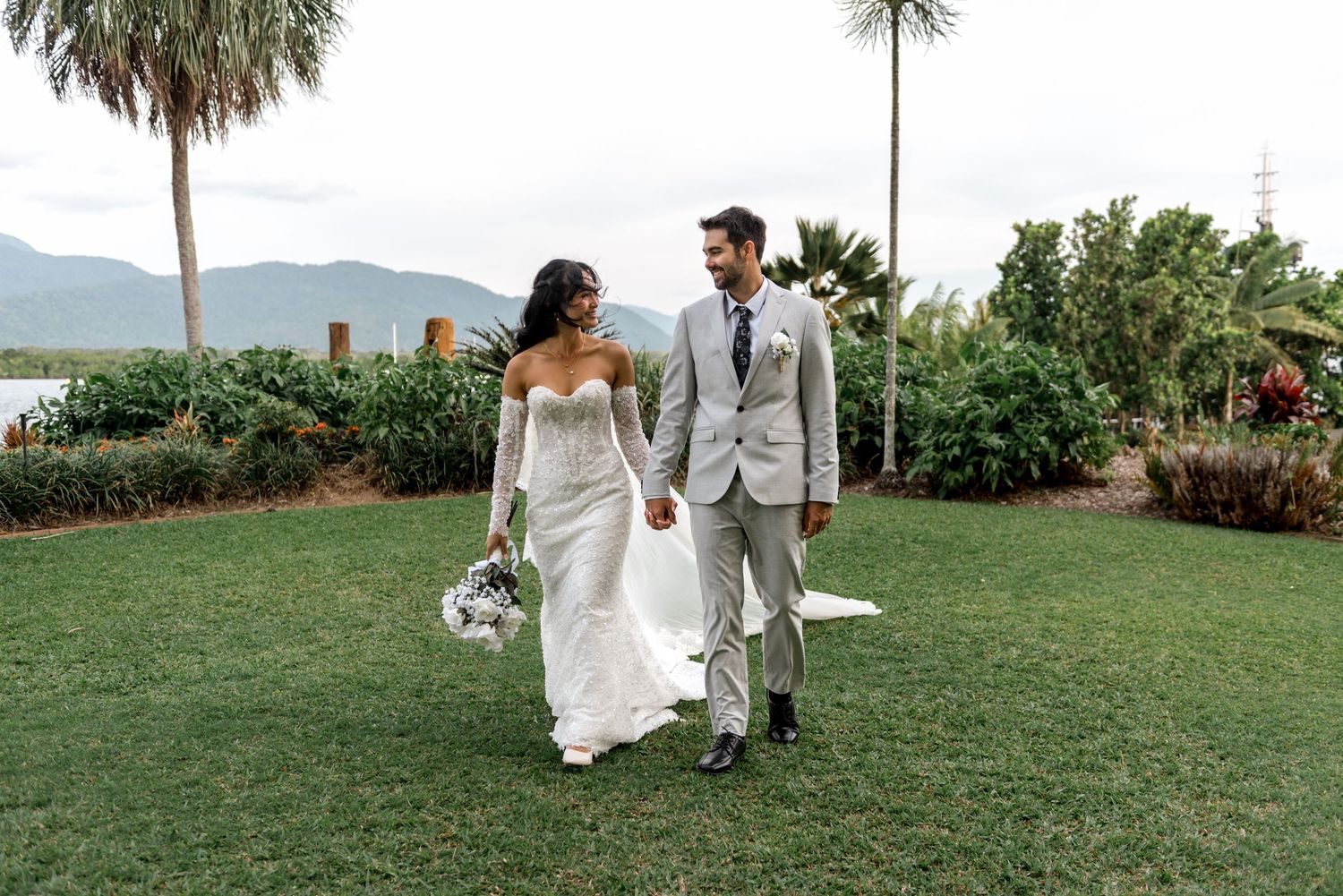 Smiling bride and groom walk hand-in-hand across green lawn with palm trees and mountains behind.