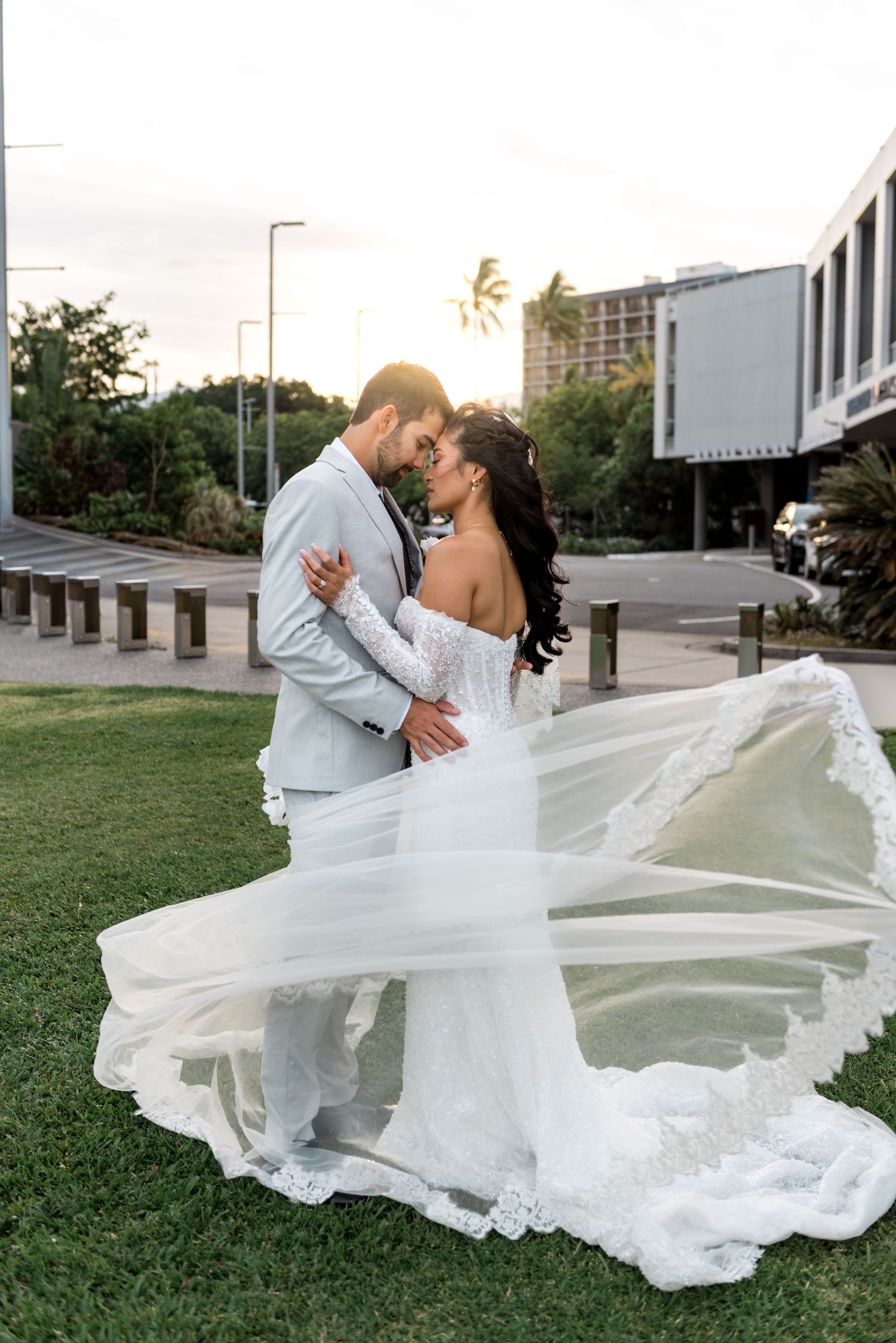 Bride's lace veil flows dramatically in the wind as couple shares a romantic sunset moment outdoors.
