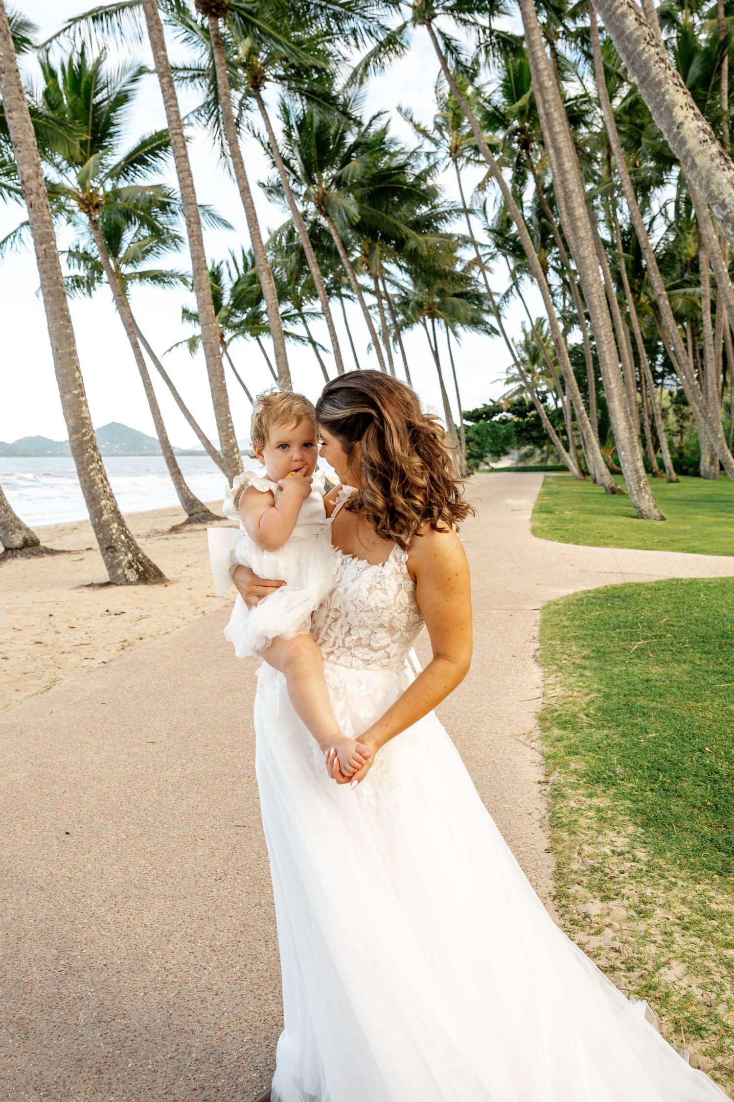 Bride holding young child in white dress along a palm-lined tropical beach path.