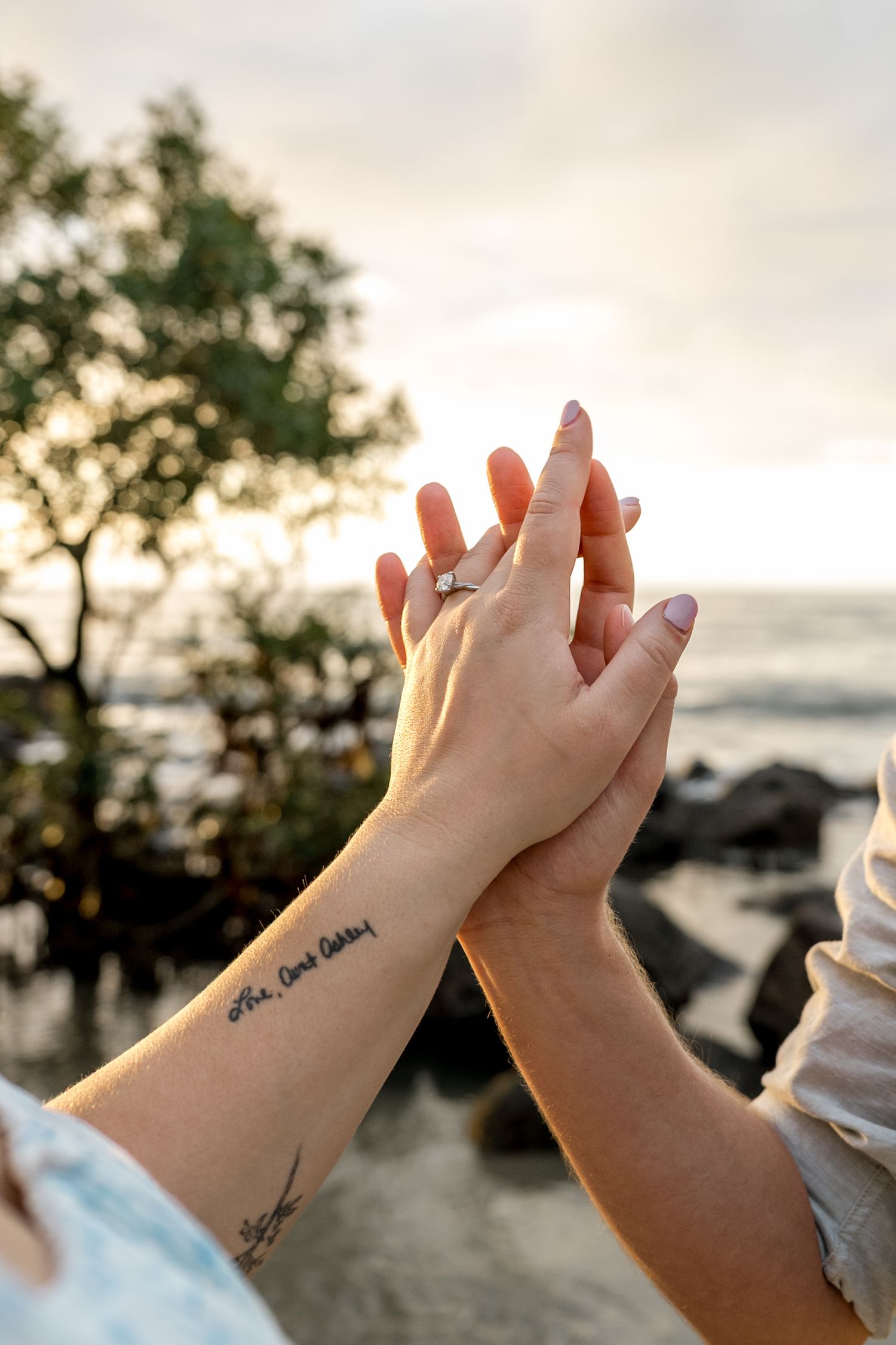 Close-up of couple holding hands on beach, engagement ring visible, tattoo on wrist, sunset backdrop at Four Mile Beach, Port Douglas.
