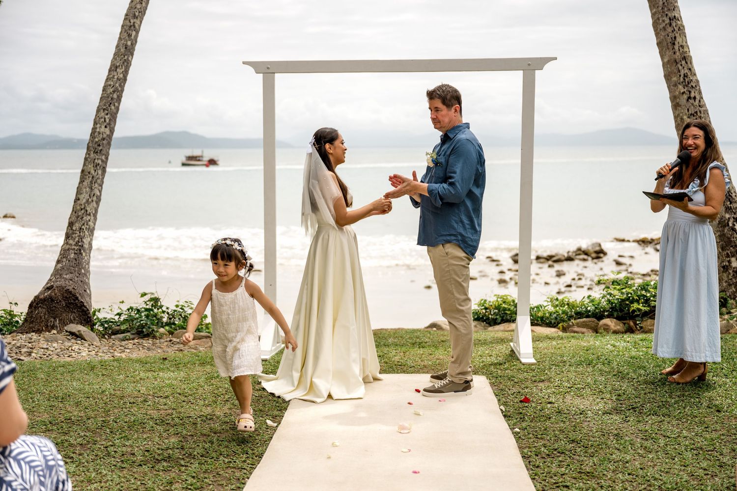 Bride and groom exchange vows at white arch altar on tropical beach as child runs past at Little Cove, Port Douglas.