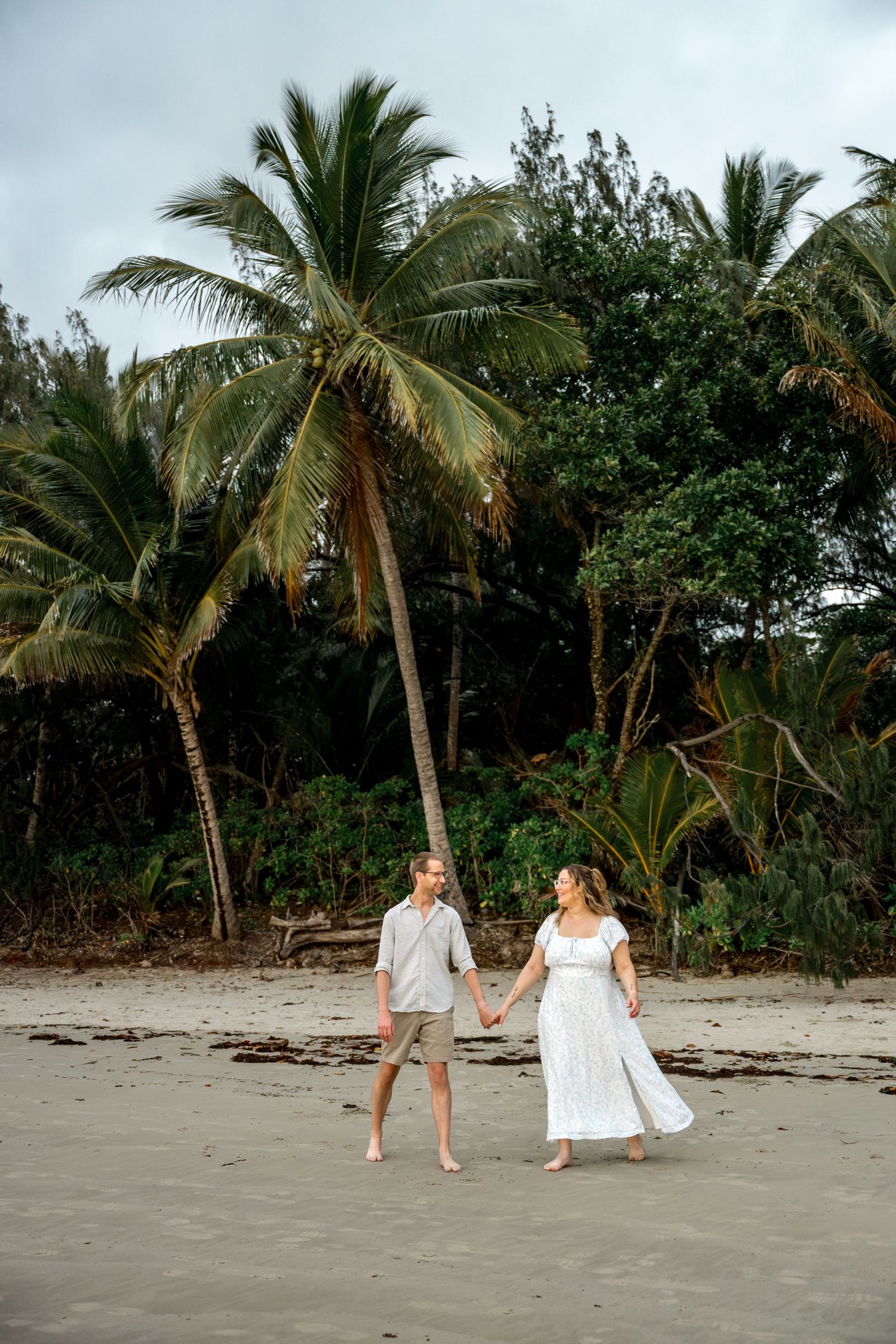 Couple holding hands and walking barefoot along sandy tropical beach with tall palm trees behind them at Four Mile Beach, Port Douglas.
