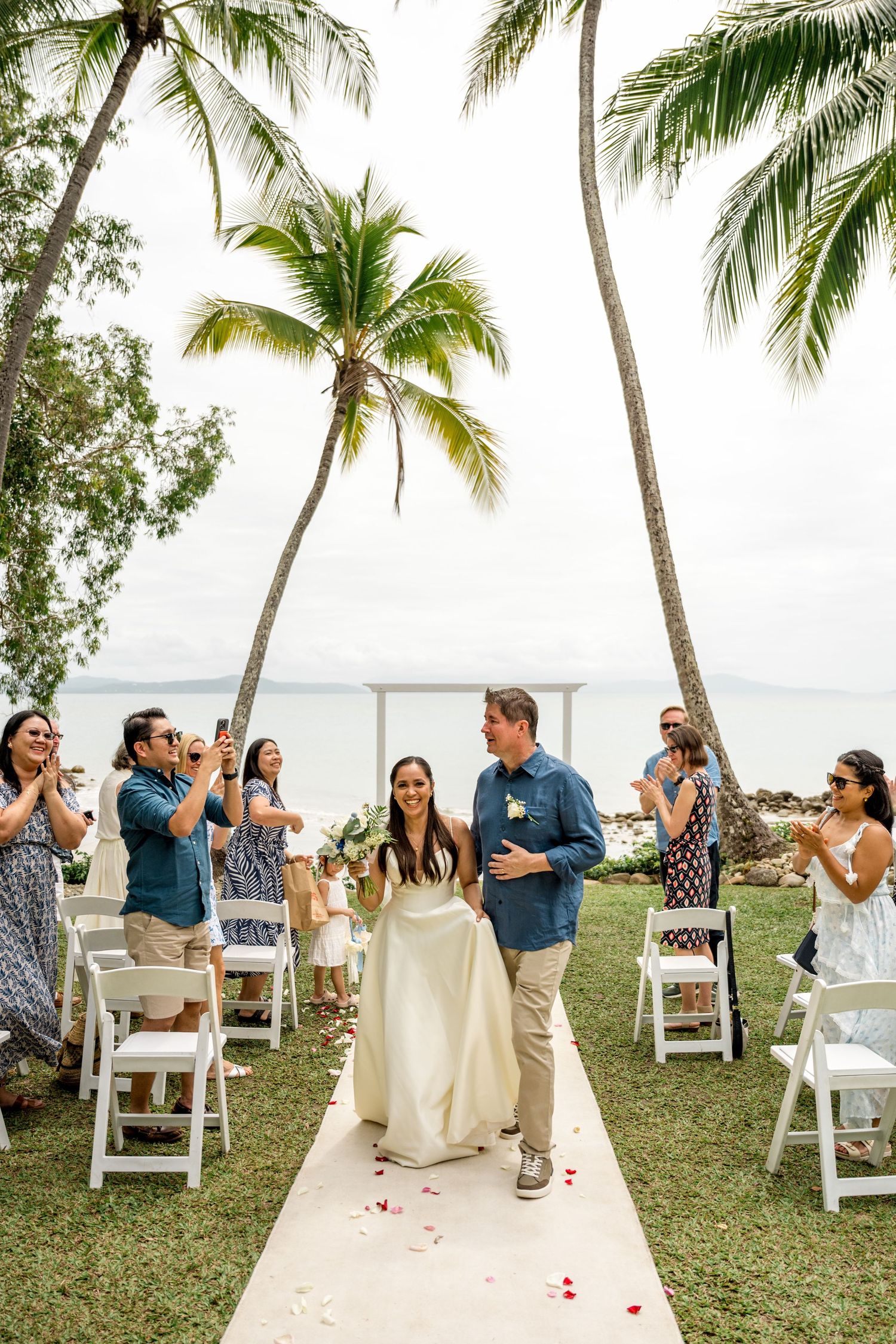 Smiling newlyweds walking down the aisle at a beachside wedding ceremony with palm trees at Little Cove, Port Douglas.