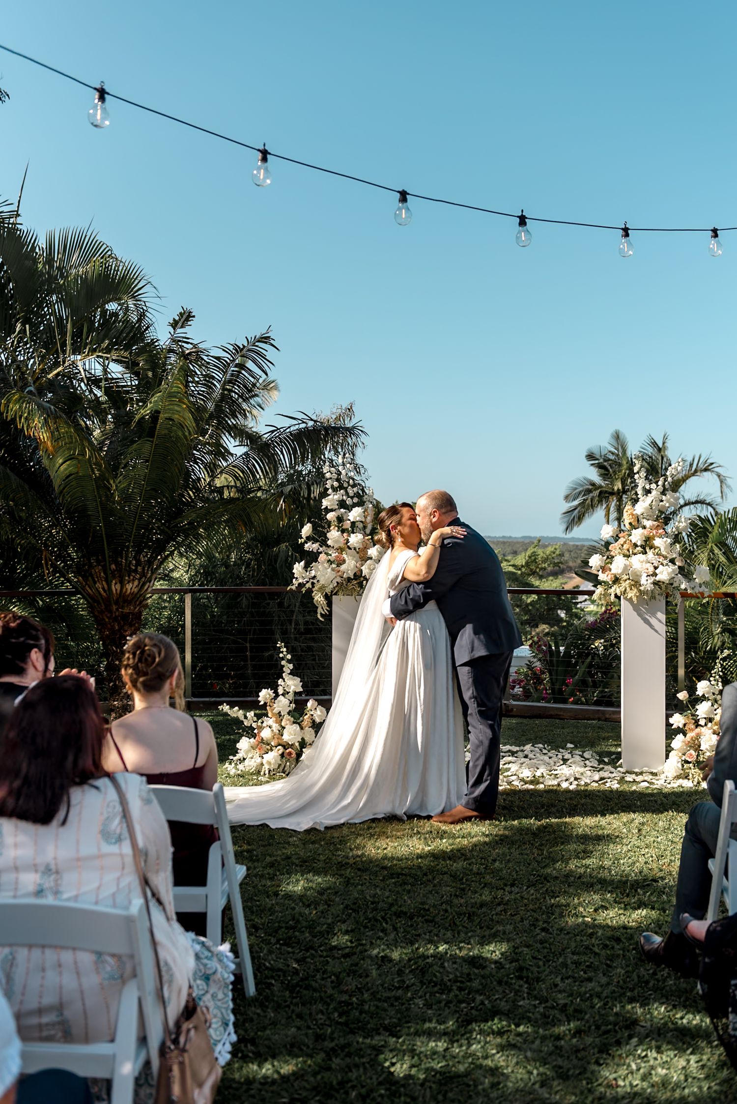 Bride and groom kissing at tropical outdoor wedding ceremony with string lights and floral arch.