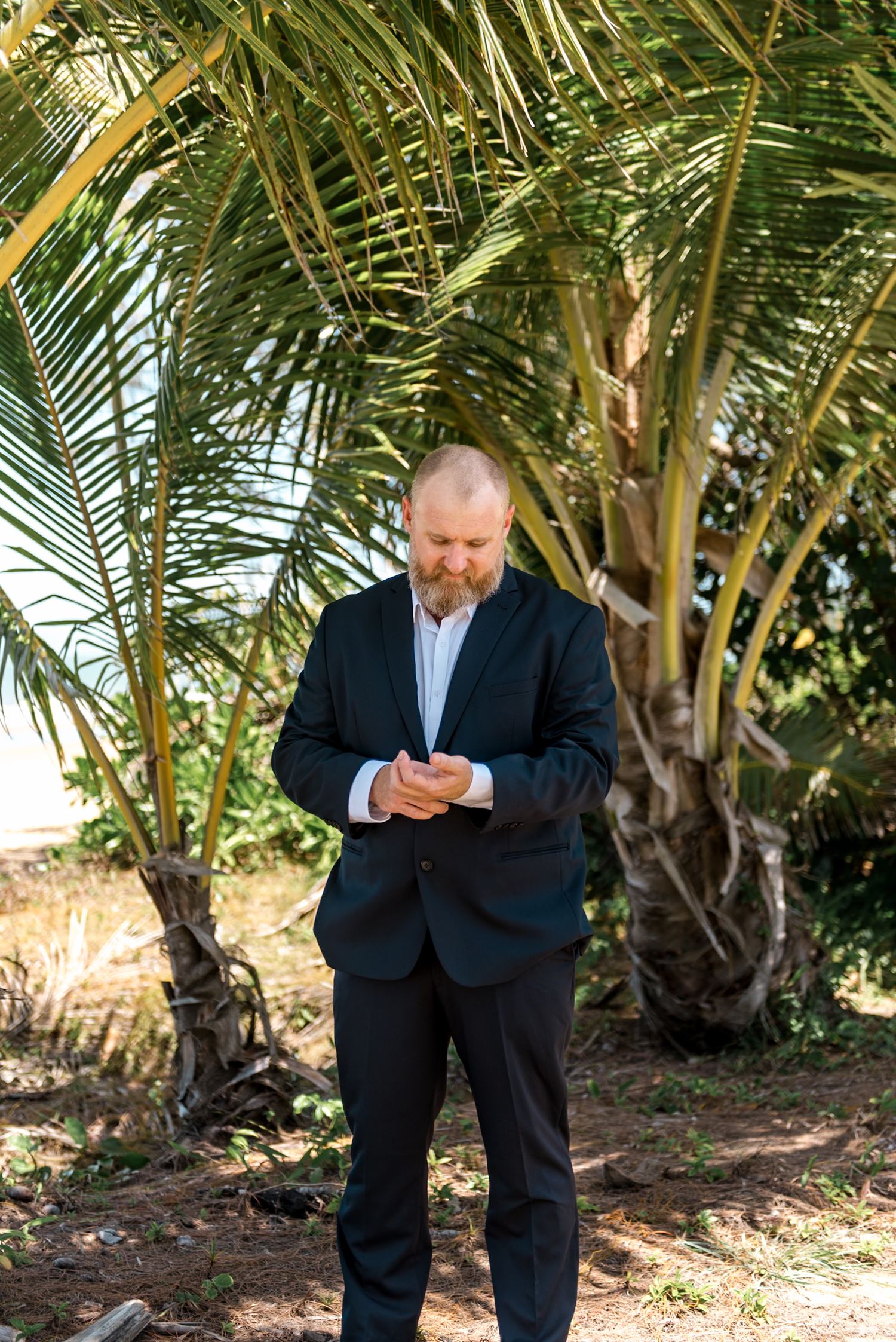 Bearded groom in dark suit looking down at wedding rings beneath tropical palm trees on sunny day.