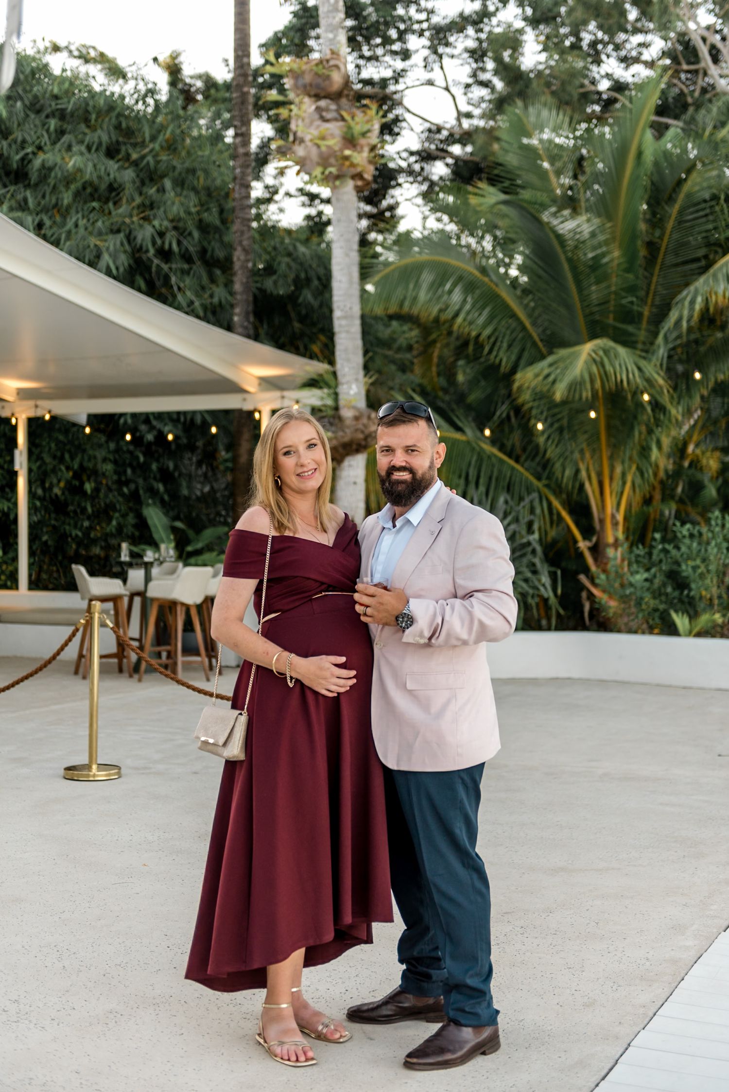 Elegant pregnant woman in burgundy dress with partner in pink blazer posing outdoors at tropical venue.
