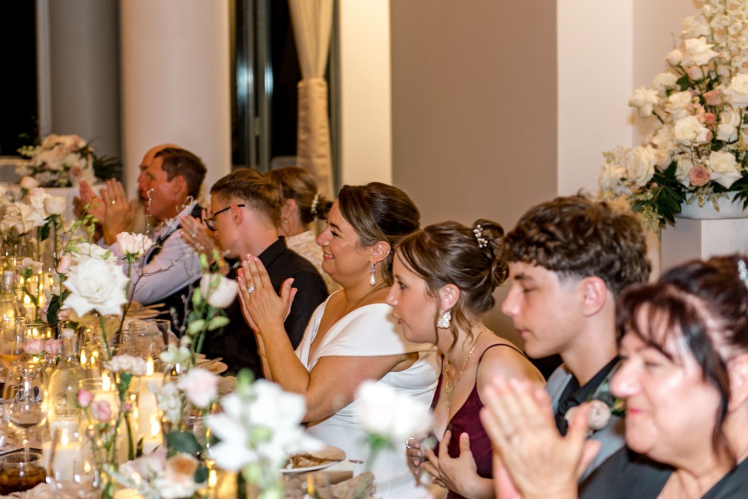 Wedding guests applauding at elegant reception with white floral centerpieces and candlelit tables.