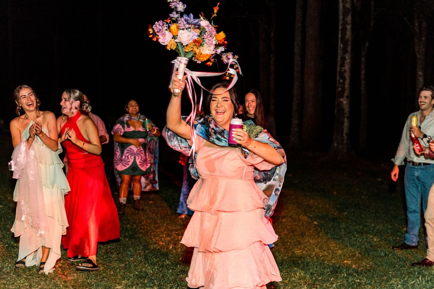 Woman in pink ruffled dress triumphantly holding a floral bouquet at a night wedding.