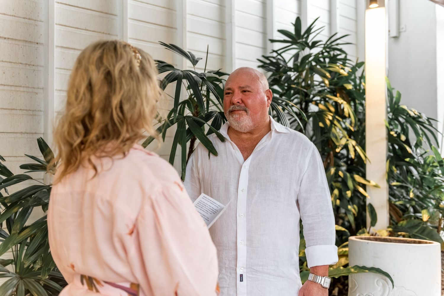 Mature groom in white linen shirt smiles at bride during outdoor vow renewal ceremony.