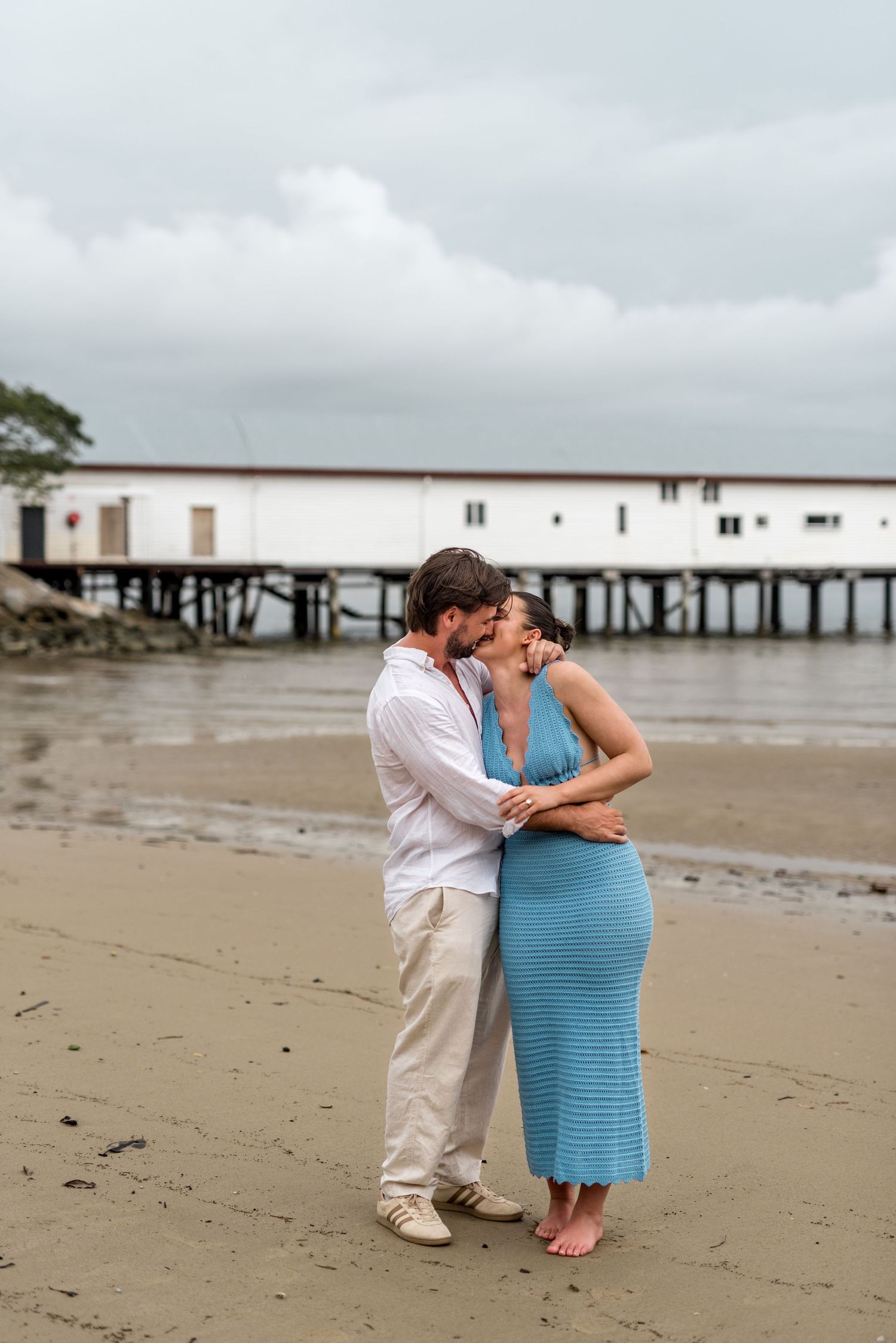 Couple sharing an intimate moment embracing on a sandy beach with a white pier building in the background.