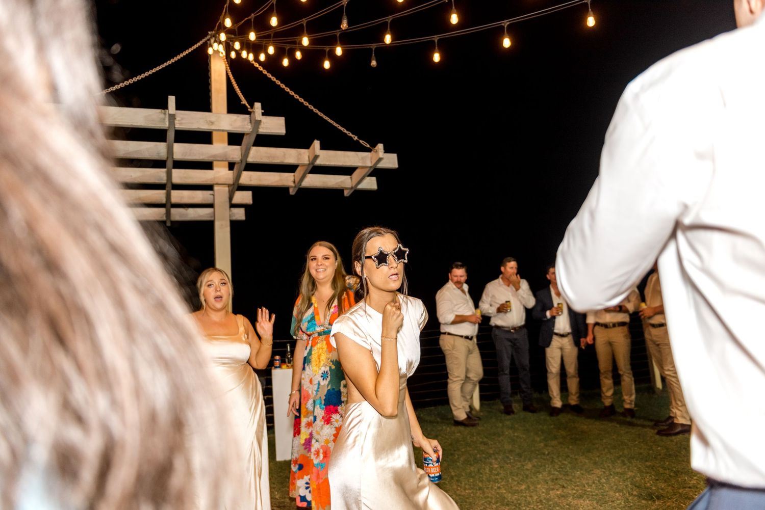 Bride in white dress with sunglasses enjoying outdoor wedding reception under string lights at night.