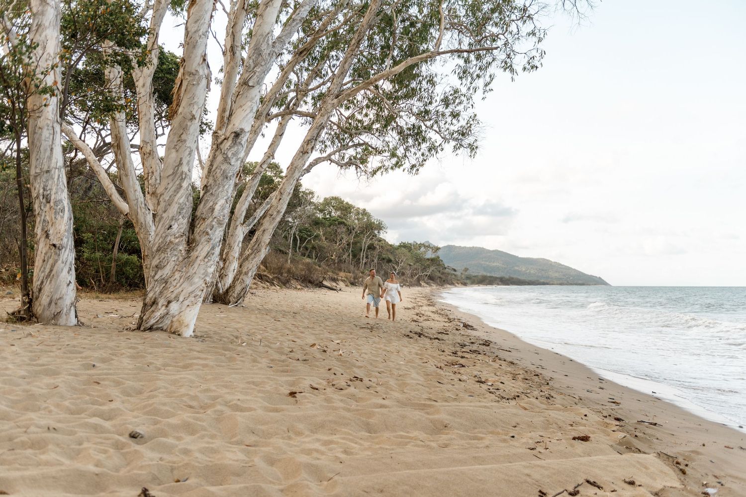 Couple strolling hand in hand along a serene sandy beach lined with white paperbark trees.