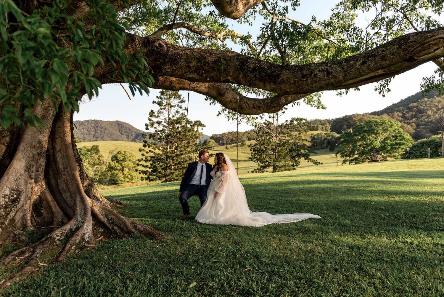 Bride and groom sit together under a sprawling tree on lush green grass at golden hour.