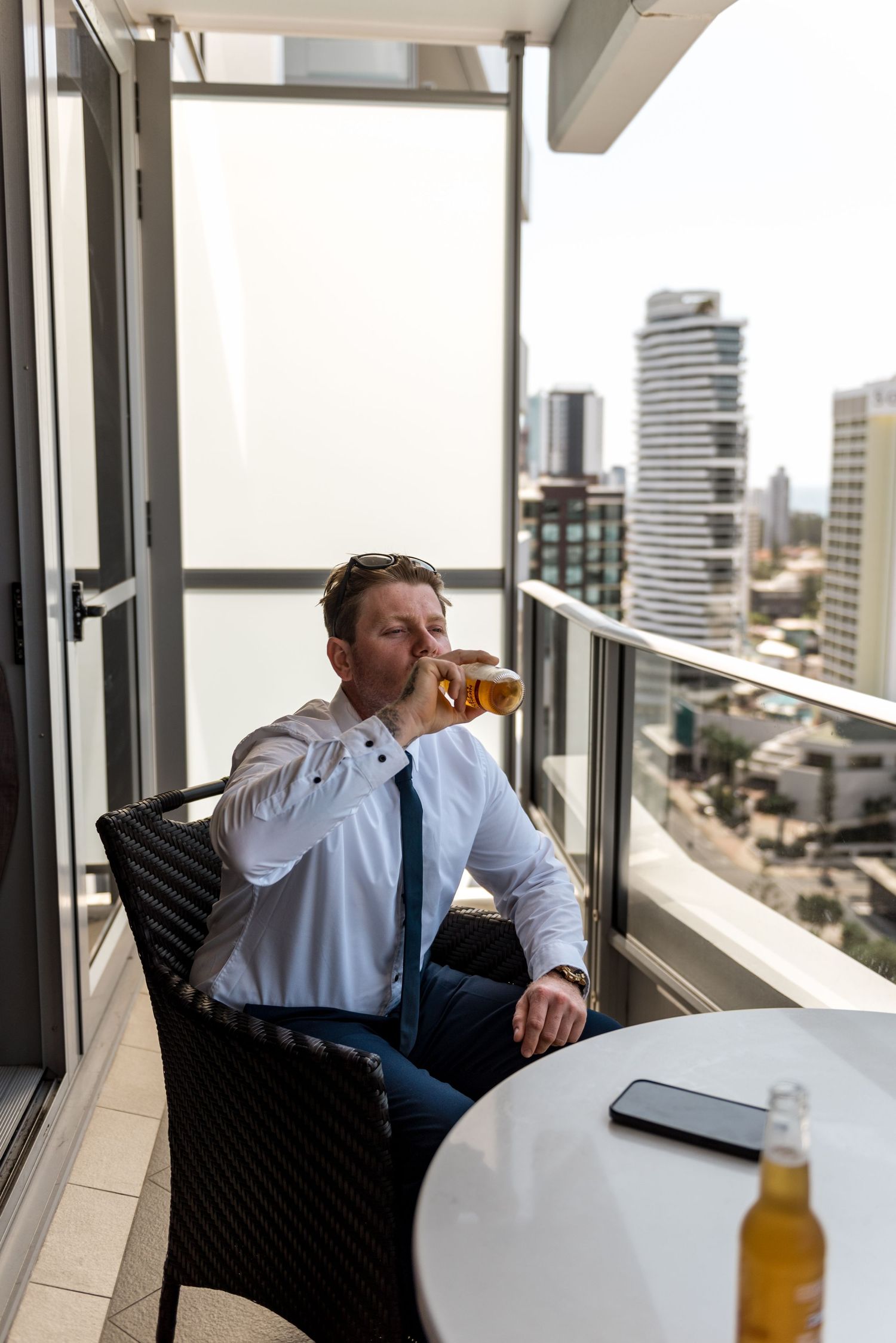 Man in white shirt drinking beer on high-rise balcony overlooking city skyline.