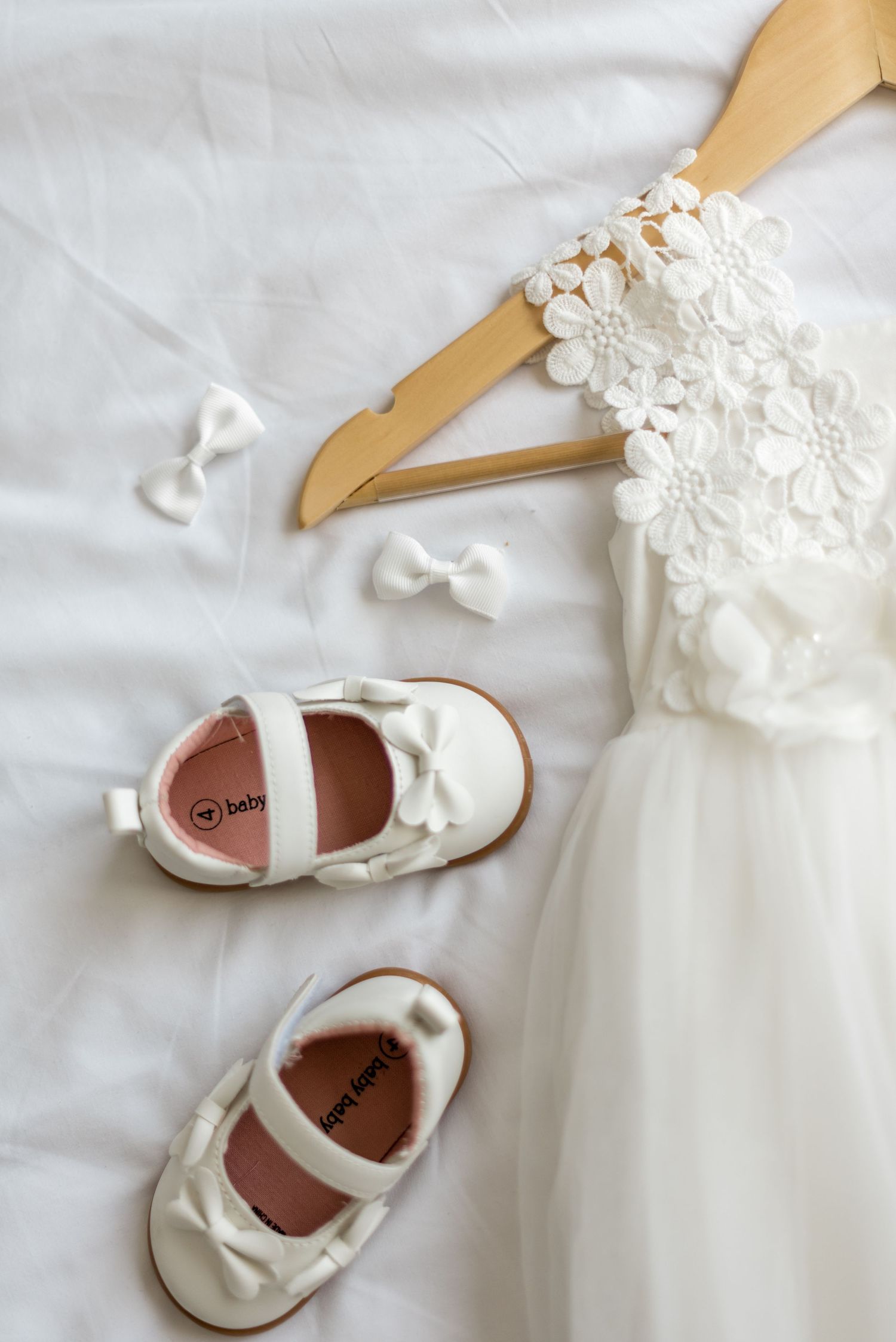 White baby Mary Jane shoes with bows beside a lace flower girl dress on a wooden hanger.