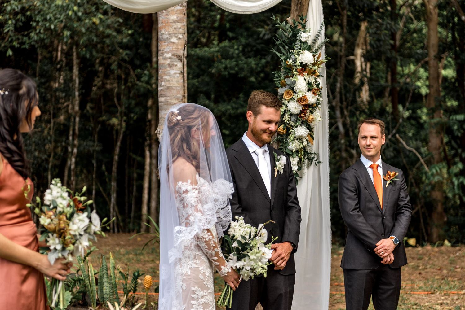 Groom smiling as bride approaches rustic floral arch at outdoor woodland wedding ceremony.