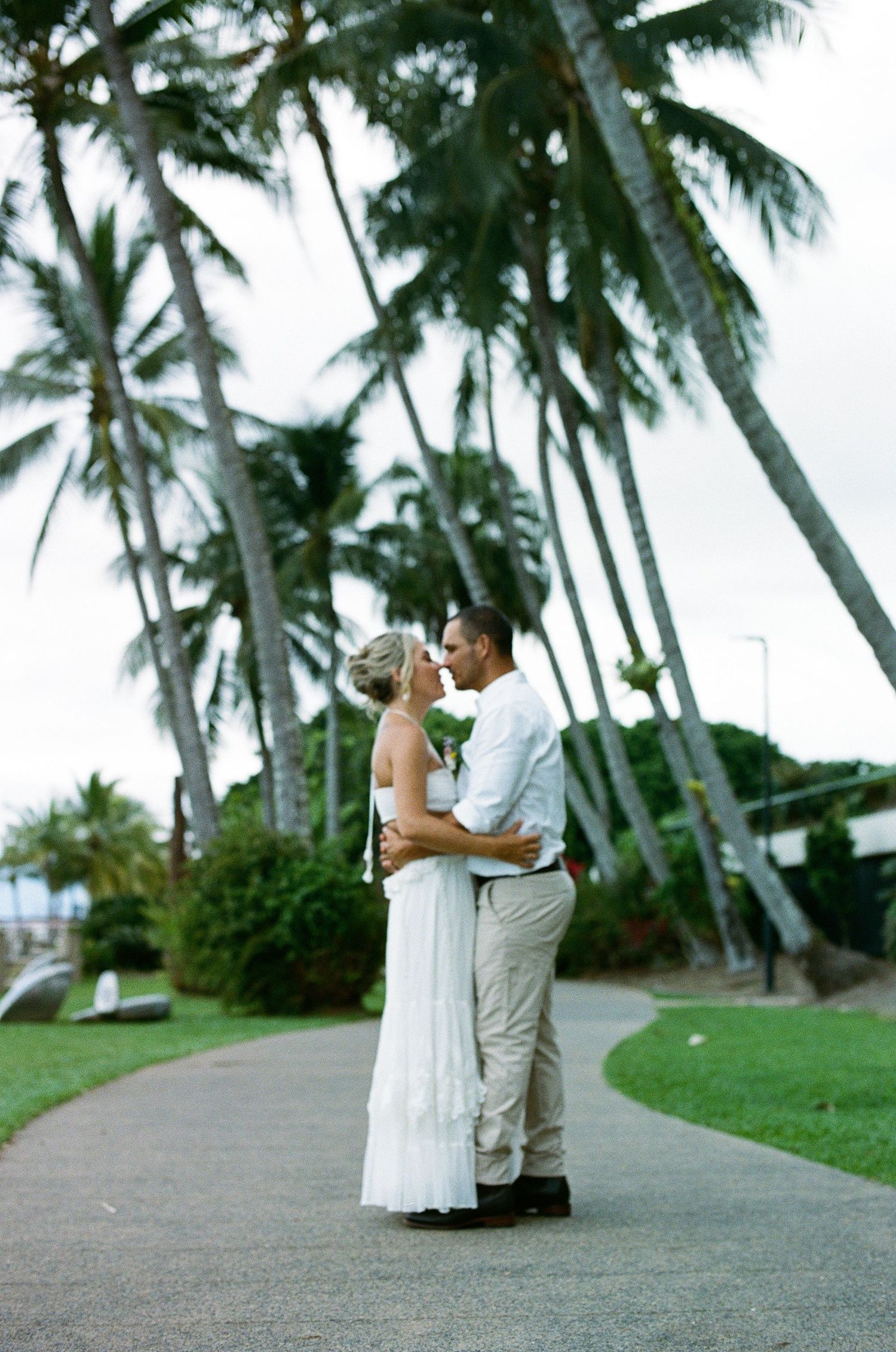 Bride and groom sharing a romantic kiss on a palm tree-lined pathway at a tropical wedding.