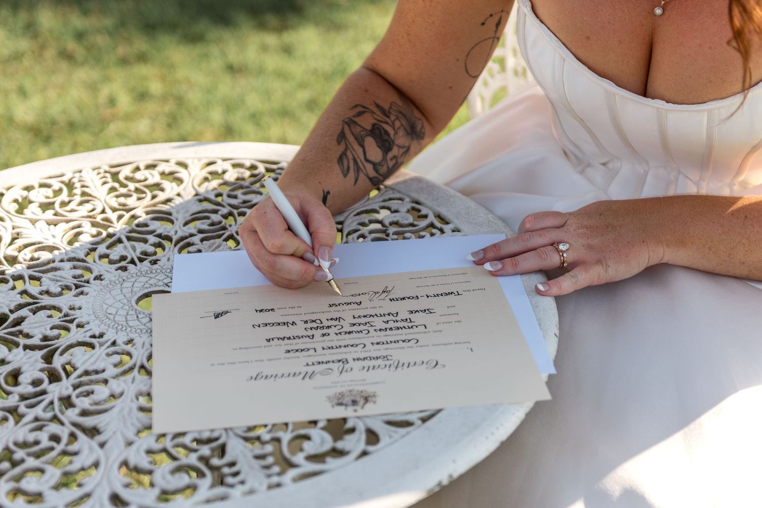 Bride with tattoed arm signs wedding certificate on ornate white table during outdoor ceremony.