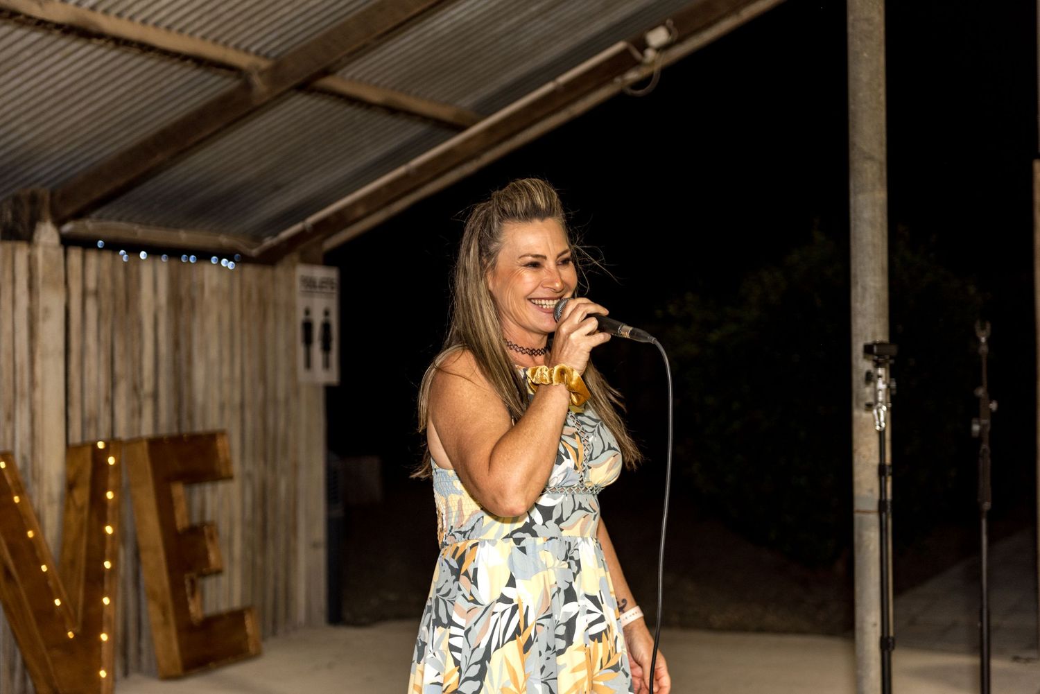 Woman in floral dress performs at outdoor wedding reception while singing into a microphone at night.