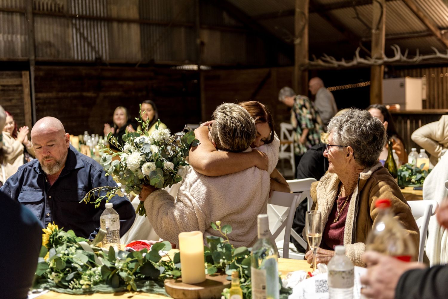 Wedding guest embraces bride with white floral bouquet at decorated barn reception dinner table.
