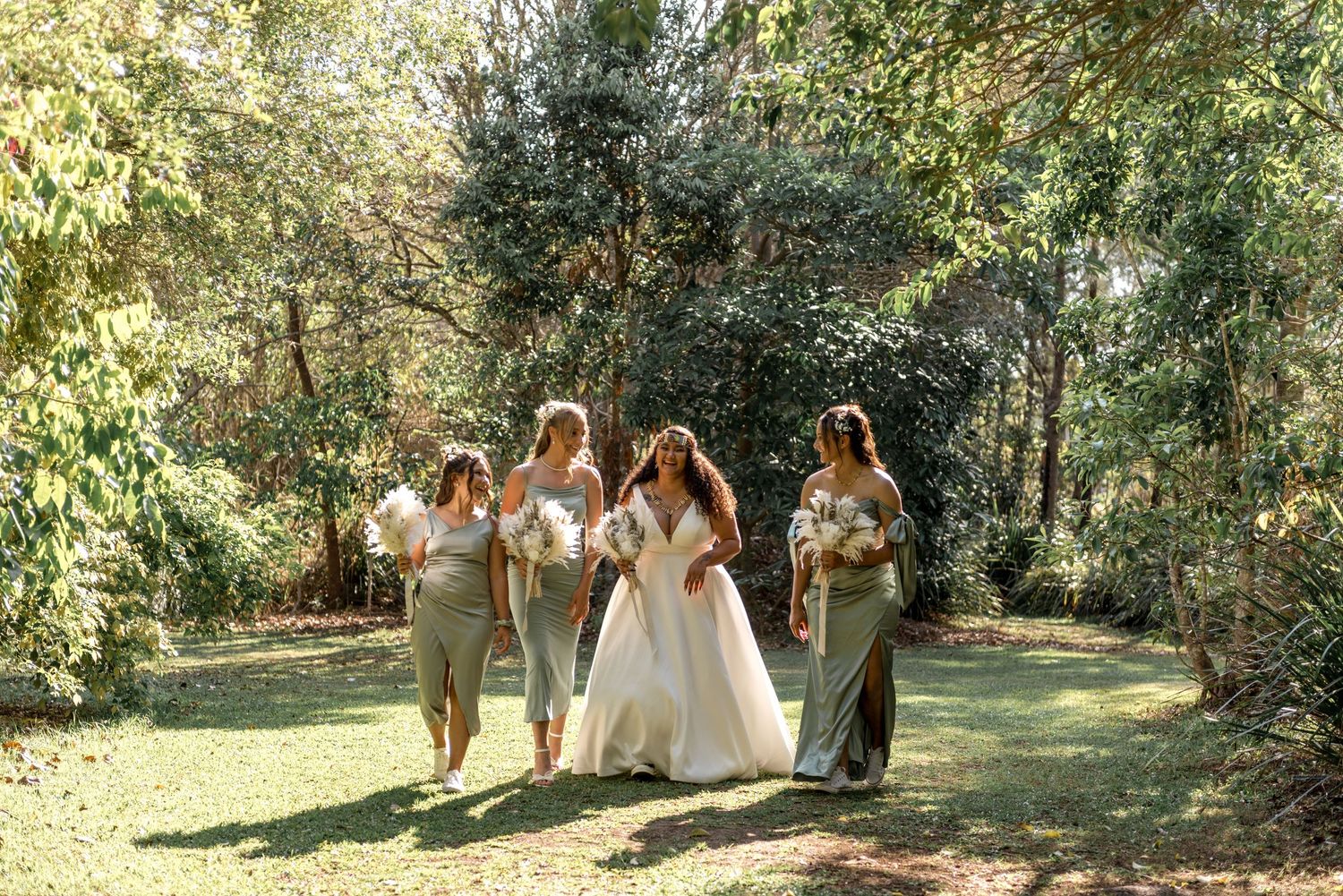 A bride in a white gown walks with four bridesmaids in sage green dresses through a garden.