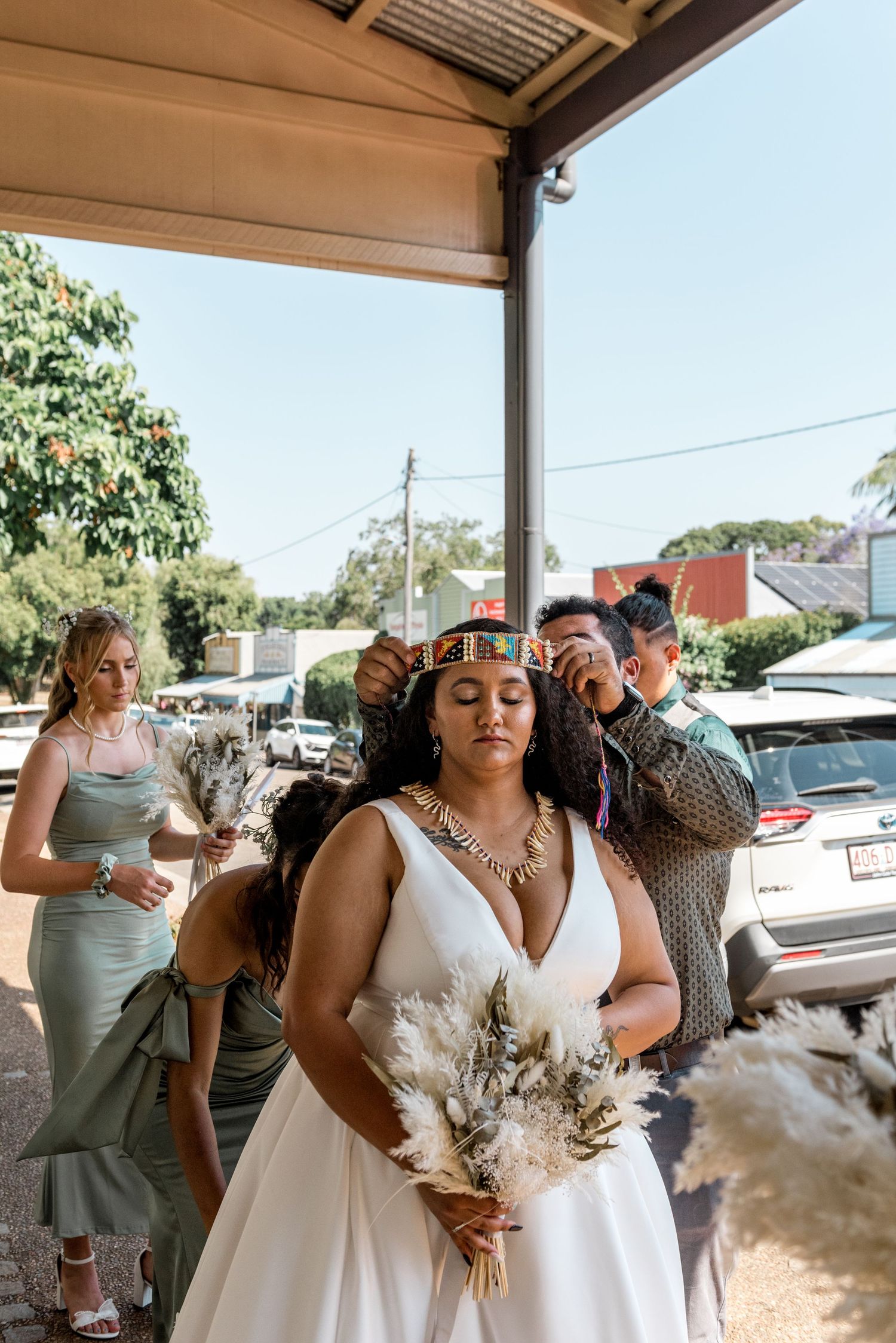Bride in white gown having traditional crown placed on head outdoors.
